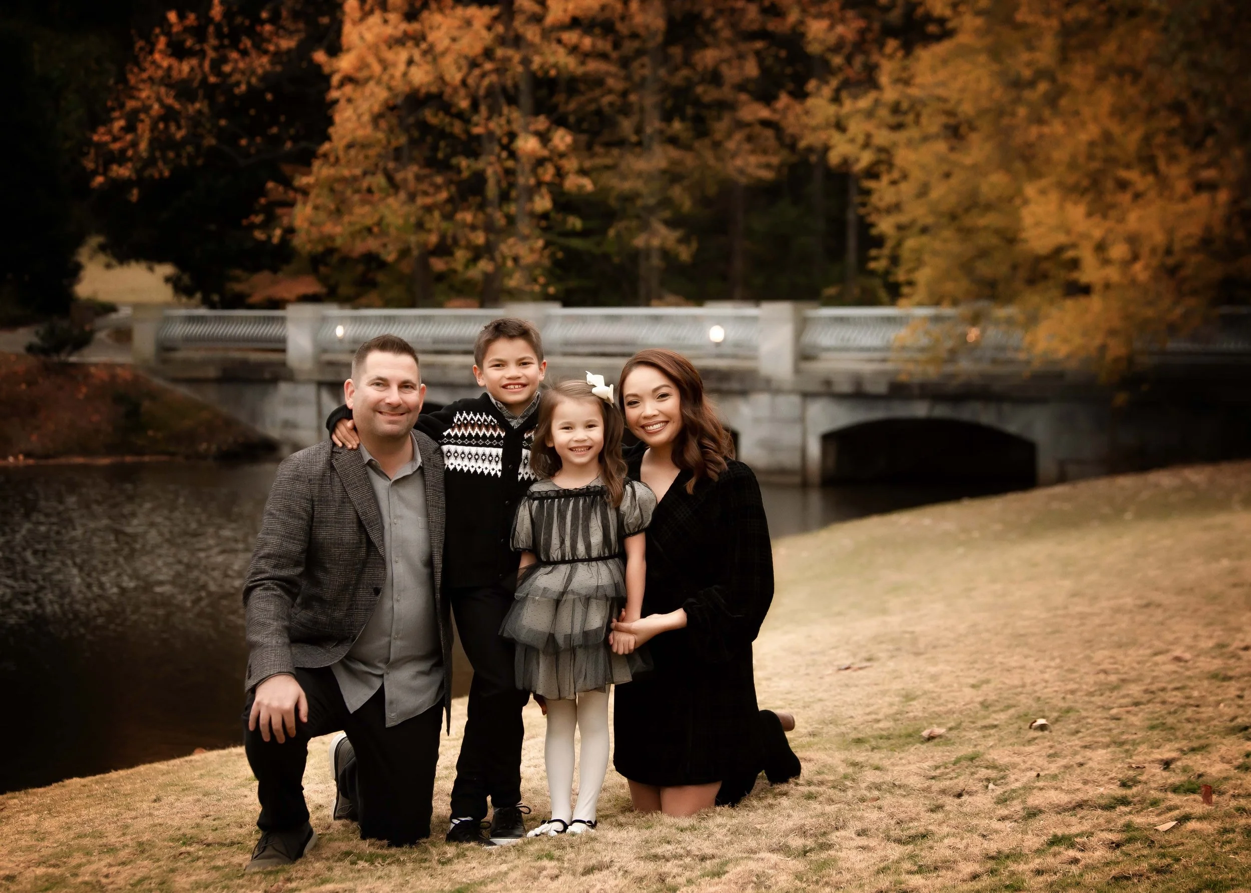A family of four, including a man, woman, a boy, and a girl, posing outdoors near a bridge over a river with fall foliage in the background.