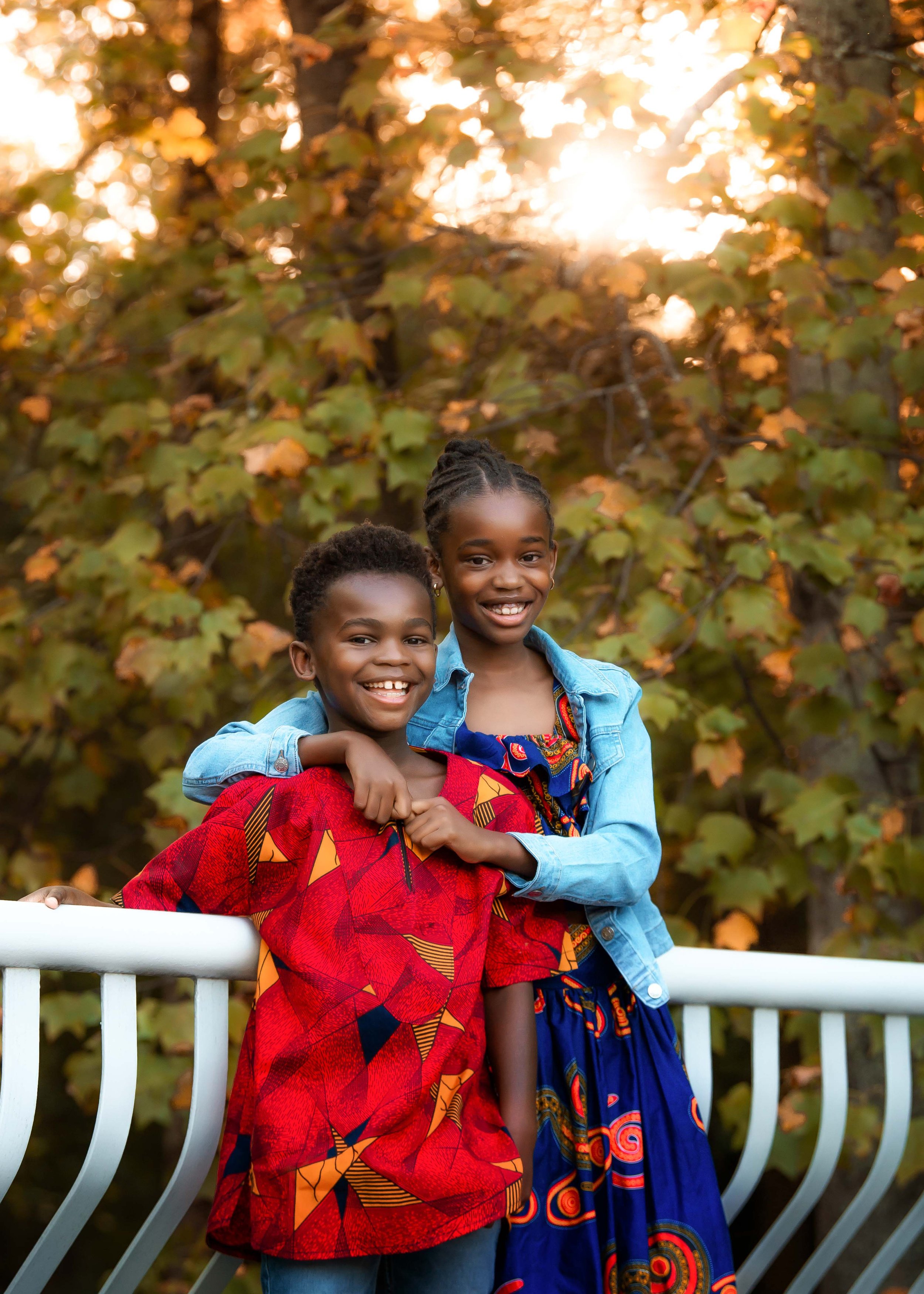 Two smiling young girls standing on a white railing outdoors, with autumn-colored leaves and trees in the background, sunlight shining through the leaves.