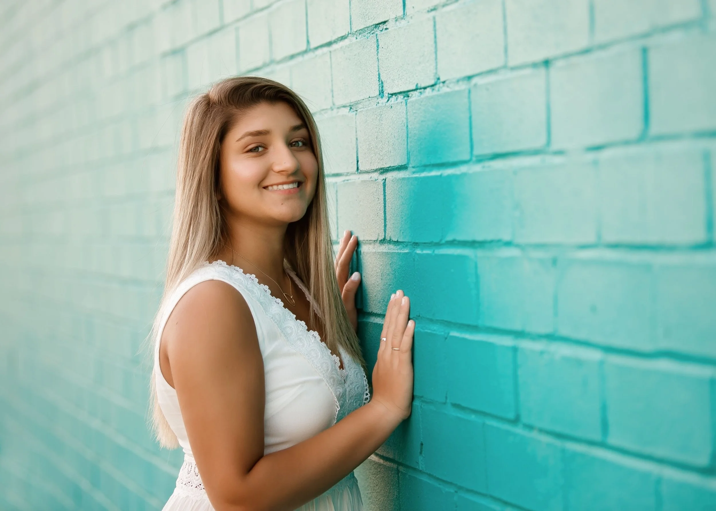 A young woman with long blonde hair smiling and leaning against a teal painted brick wall.