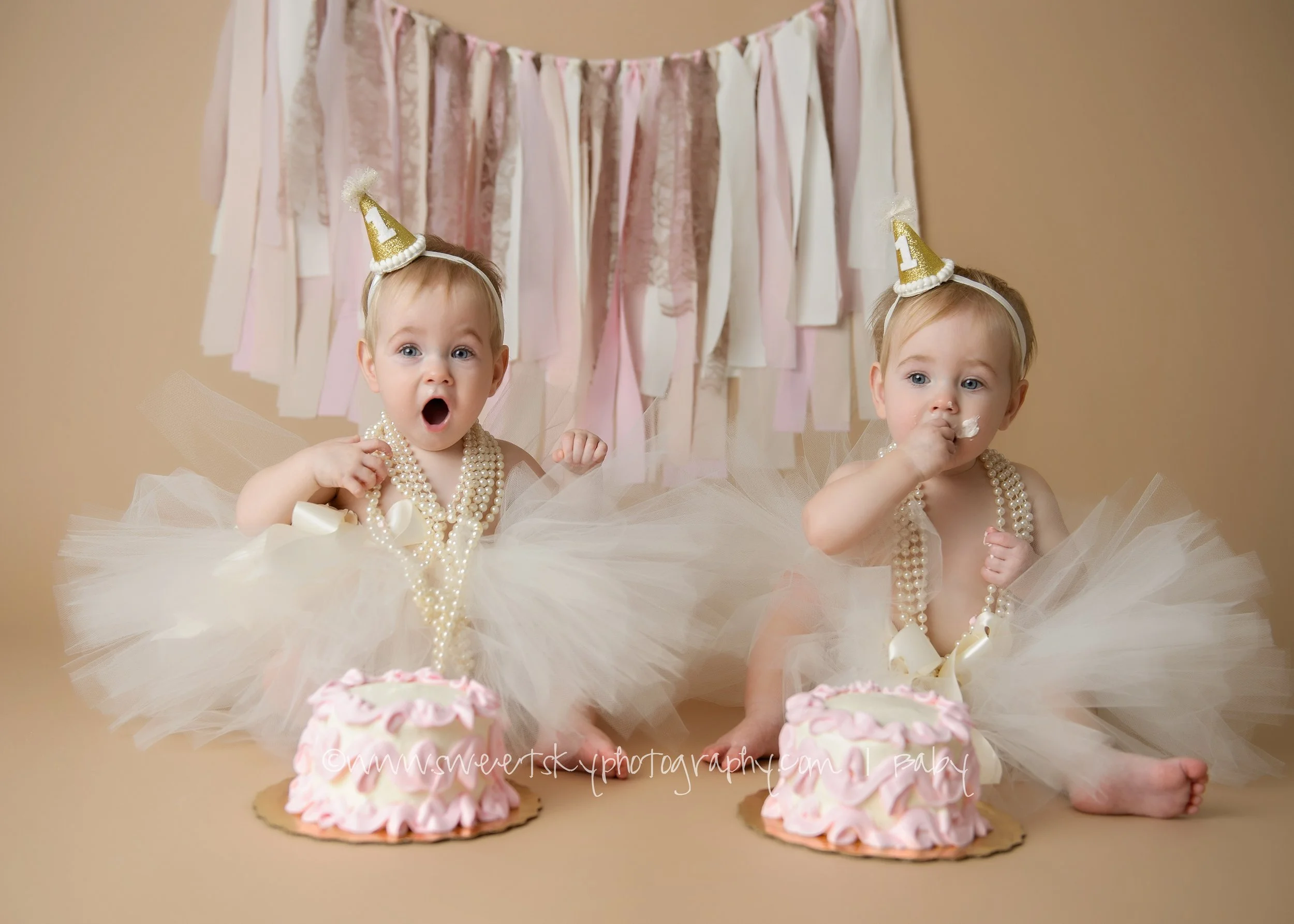 Two babies dressed in white tutu dresses with pearl necklaces, sitting in front of pink and white birthday cakes, wearing gold birthday hats with the number '1', celebrating their first birthday.