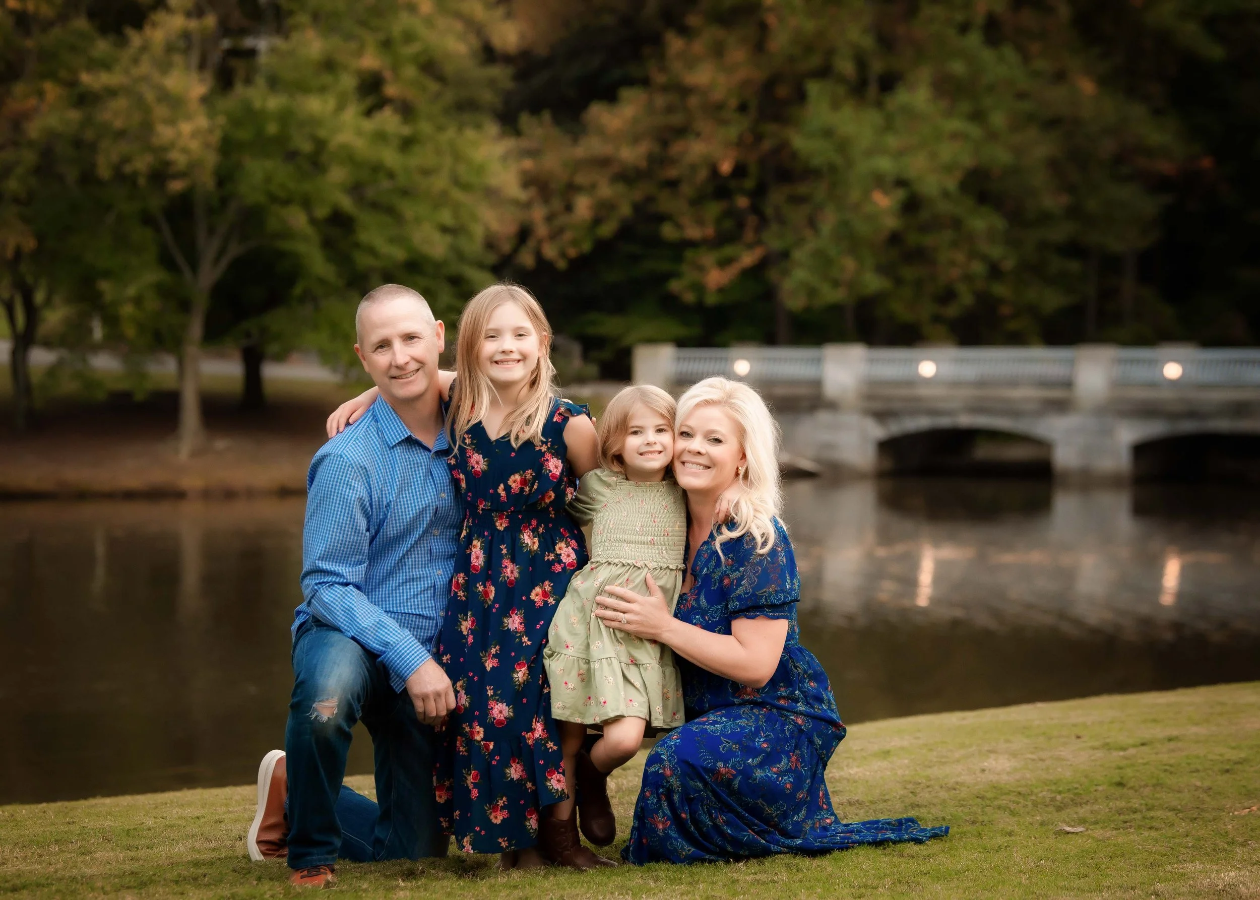 A family of four, including two children and their parents, smiling and posing together outdoors near a river with a stone bridge in the background.