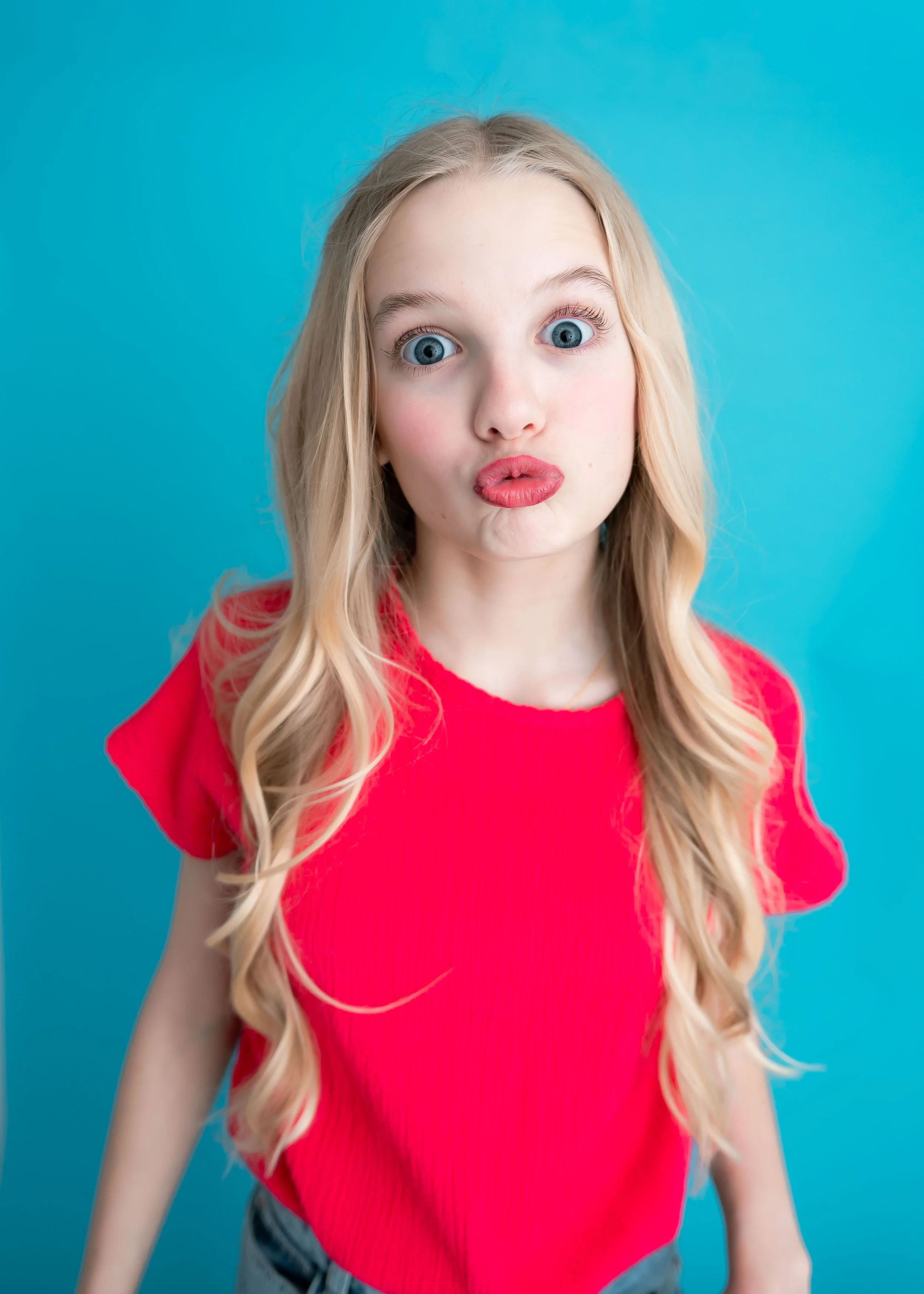 A young girl with long blonde hair and blue eyes making a funny face with puckered lips against a blue background.