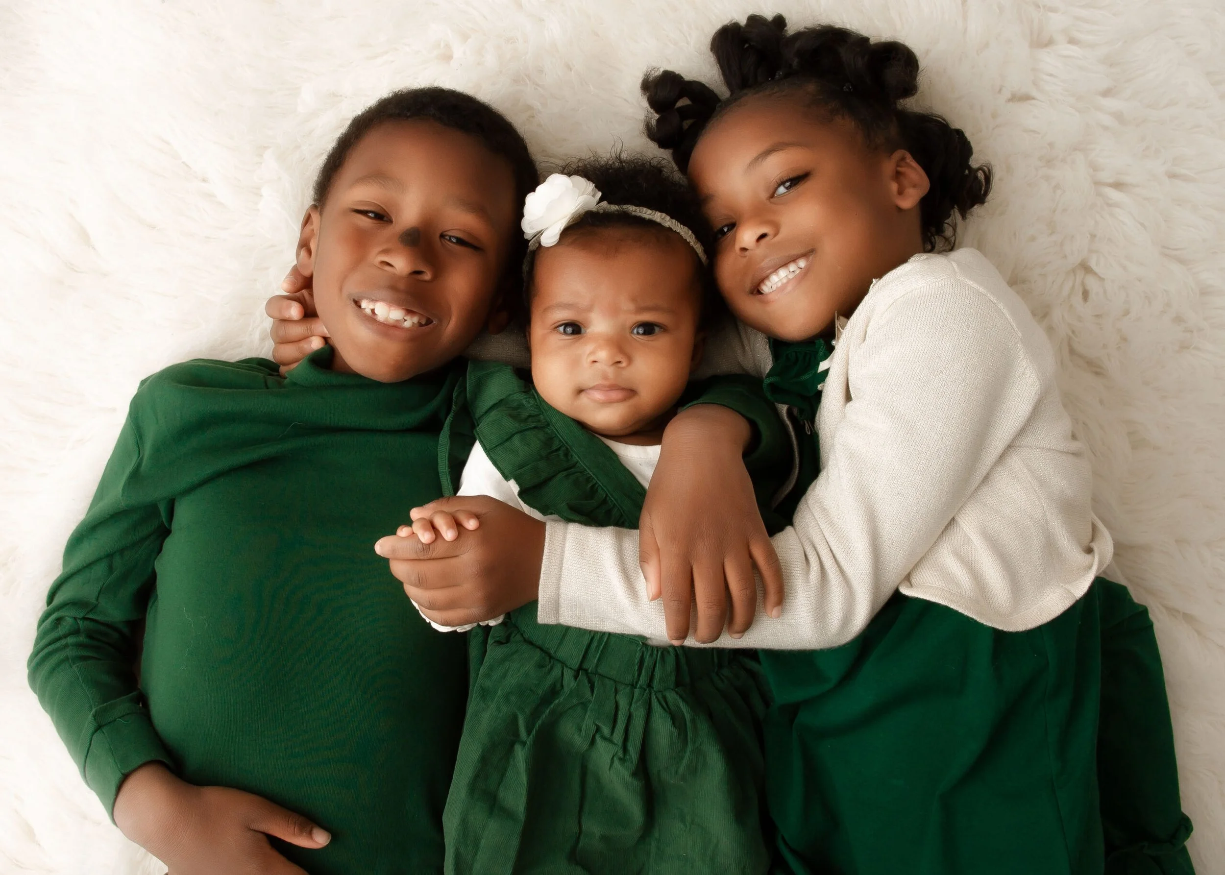 Three children, two girls and one boy, lying on a soft, white fluffy surface, wearing green and white clothing, smiling and hugging each other.