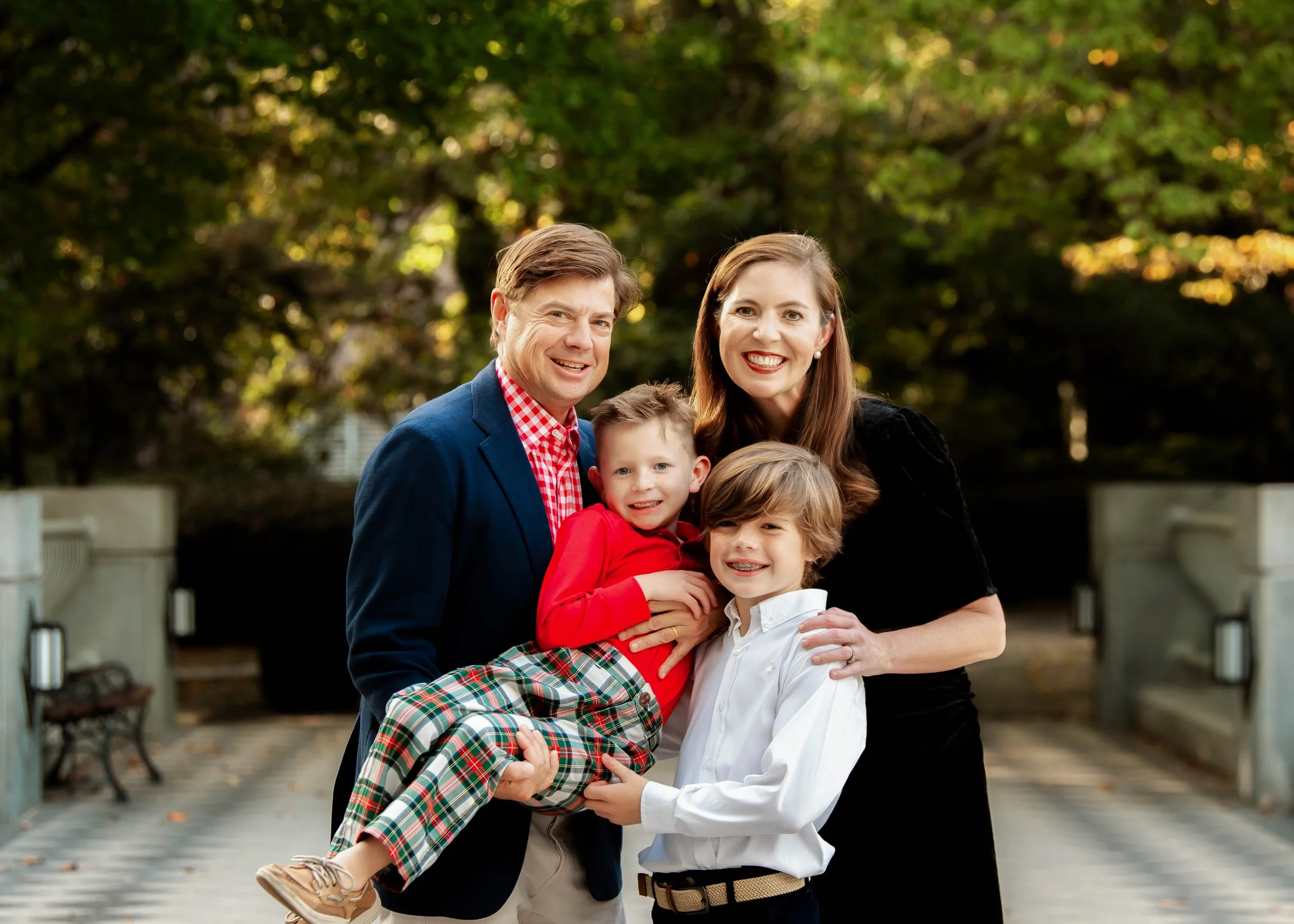 A smiling family of four outdoors during daytime, with trees and a bridge in the background. The father is holding a young boy in red plaid pants and a red shirt, while the mother and older boy in a white shirt and black pants stand beside them.