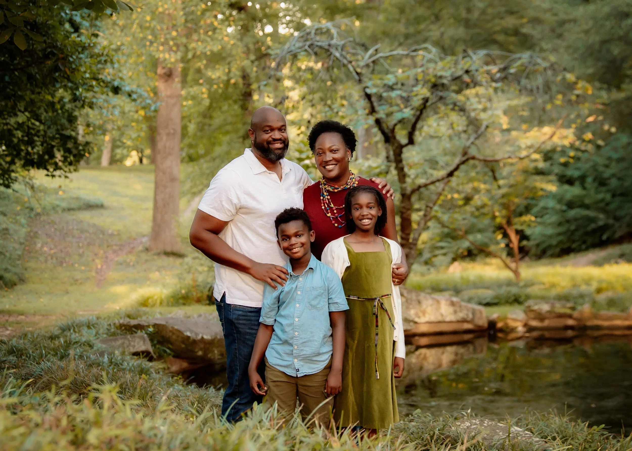 A smiling family of four standing outdoors in a park with trees and greenery, near a small pond, during daytime.