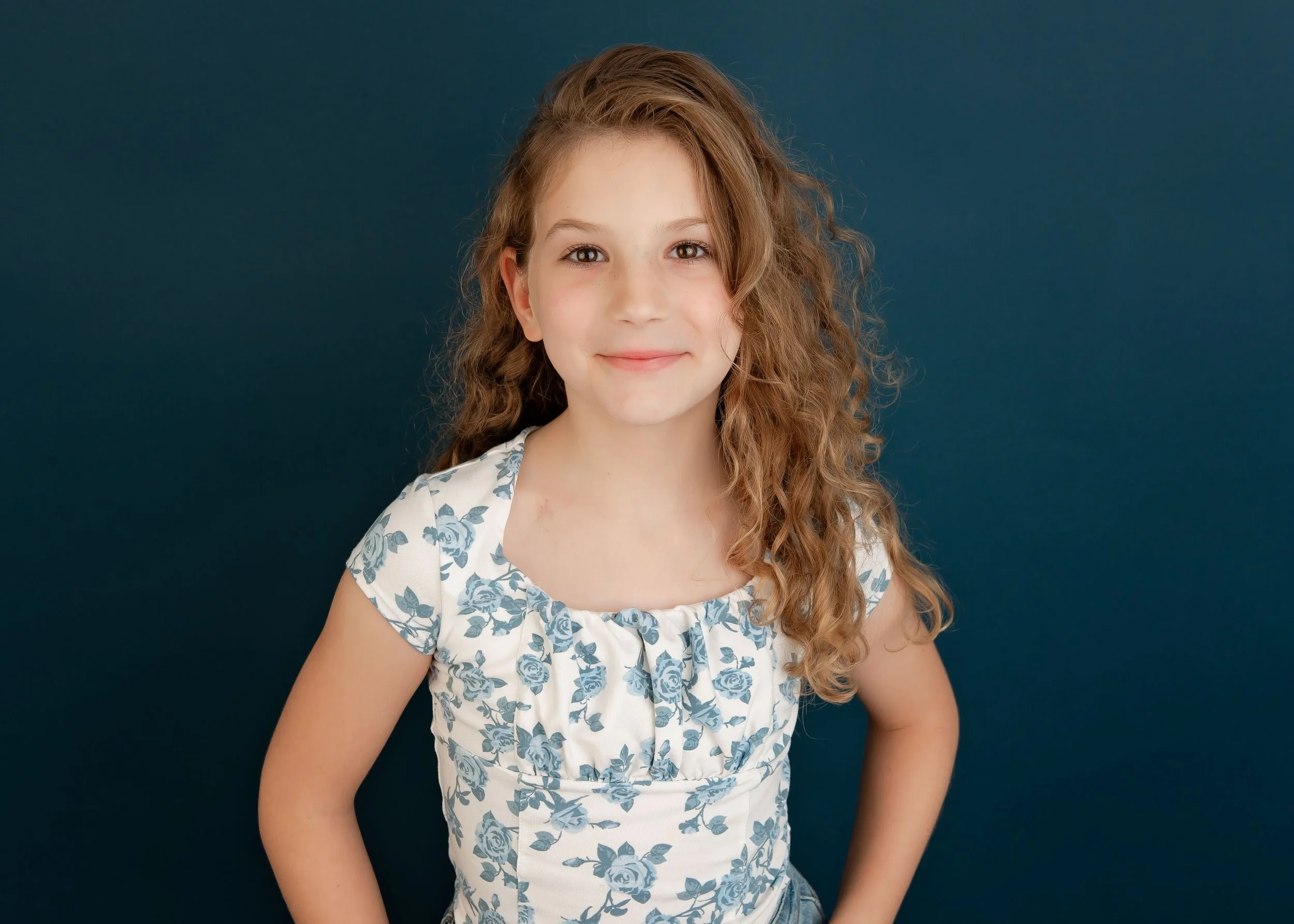 A young girl with curly brown hair and light skin wearing a short-sleeved white dress with blue floral patterns, smiling with her hands on her hips, standing against a dark blue background.
