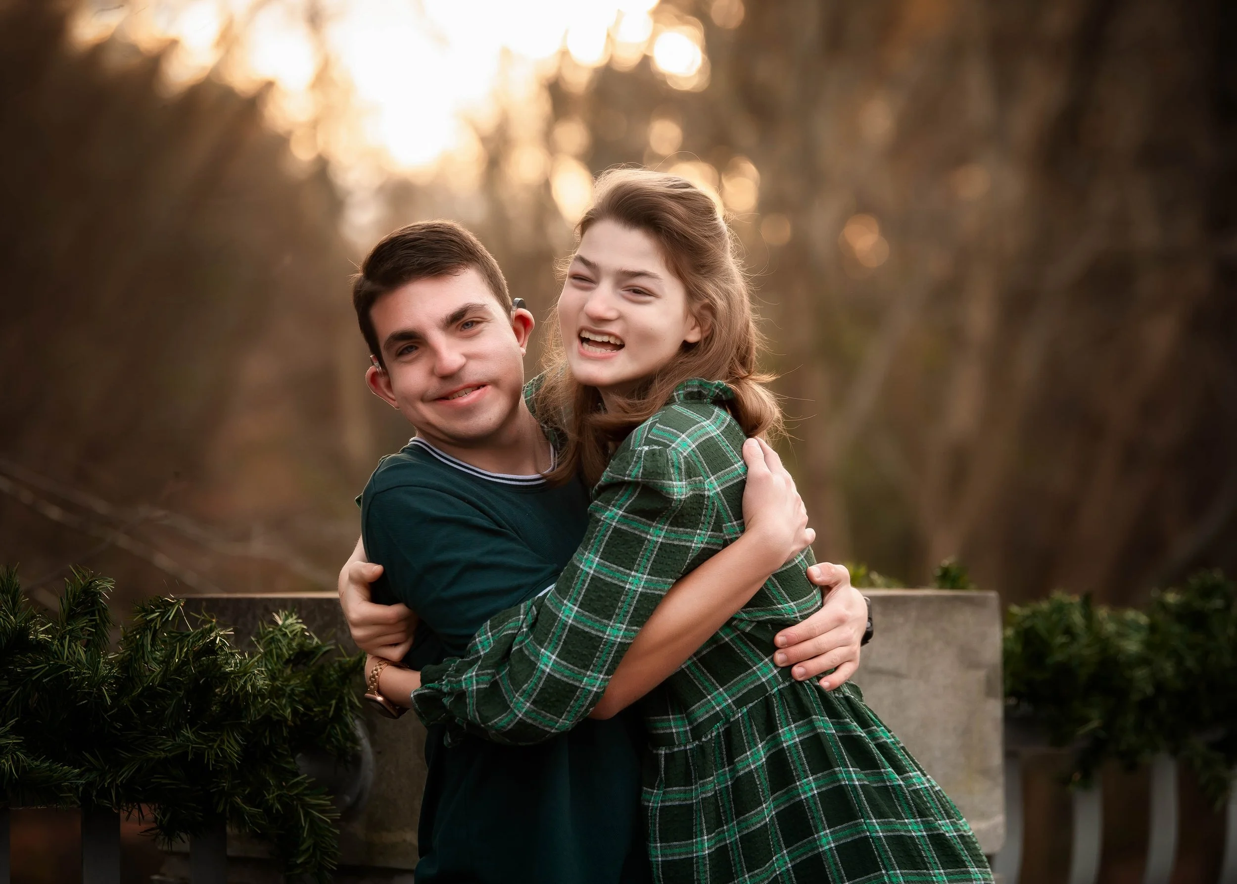 A young man and woman are hugging outdoors on a sunny day in a wooded area, both smiling and facing the camera.