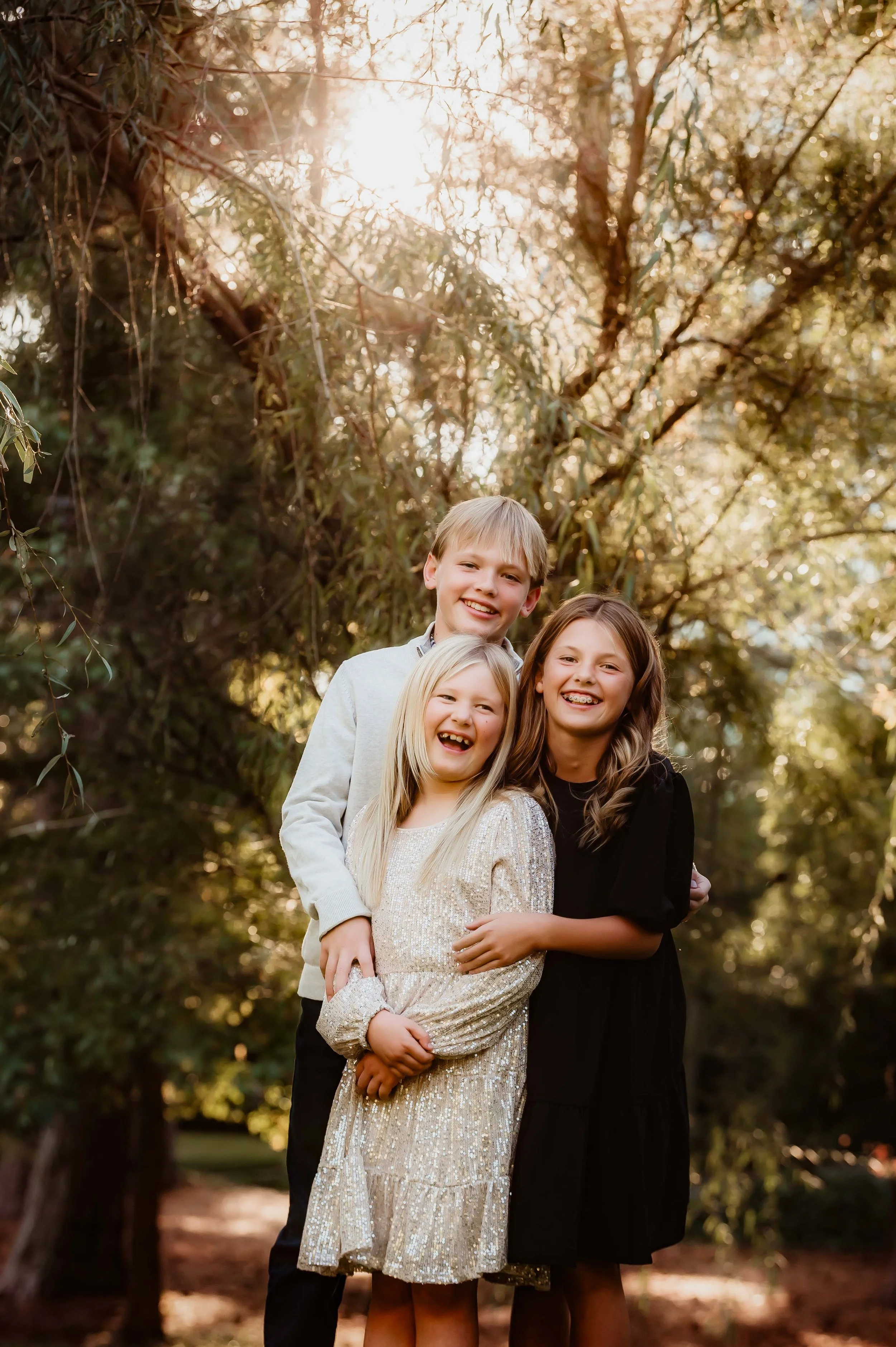 Four smiling children, two girls and two boys, standing close together outdoors during daytime with sunlight filtering through trees in the background.