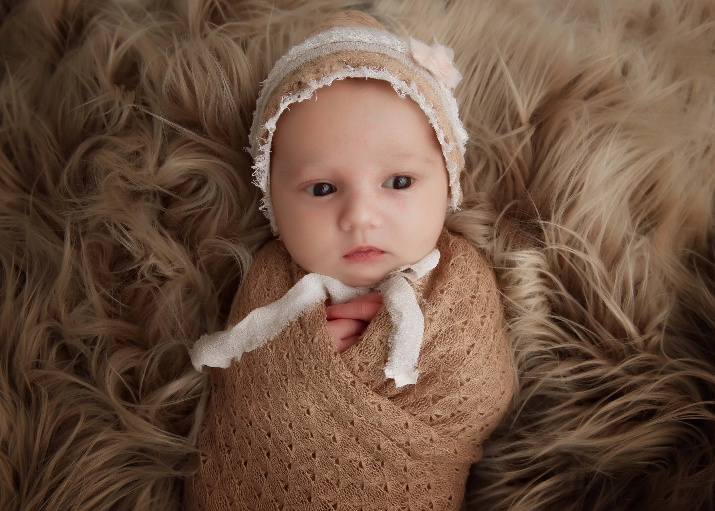 A baby with big, expressive eyes lying on a fluffy, brown fur blanket, wearing a lace bonnet and a brown lace outfit.