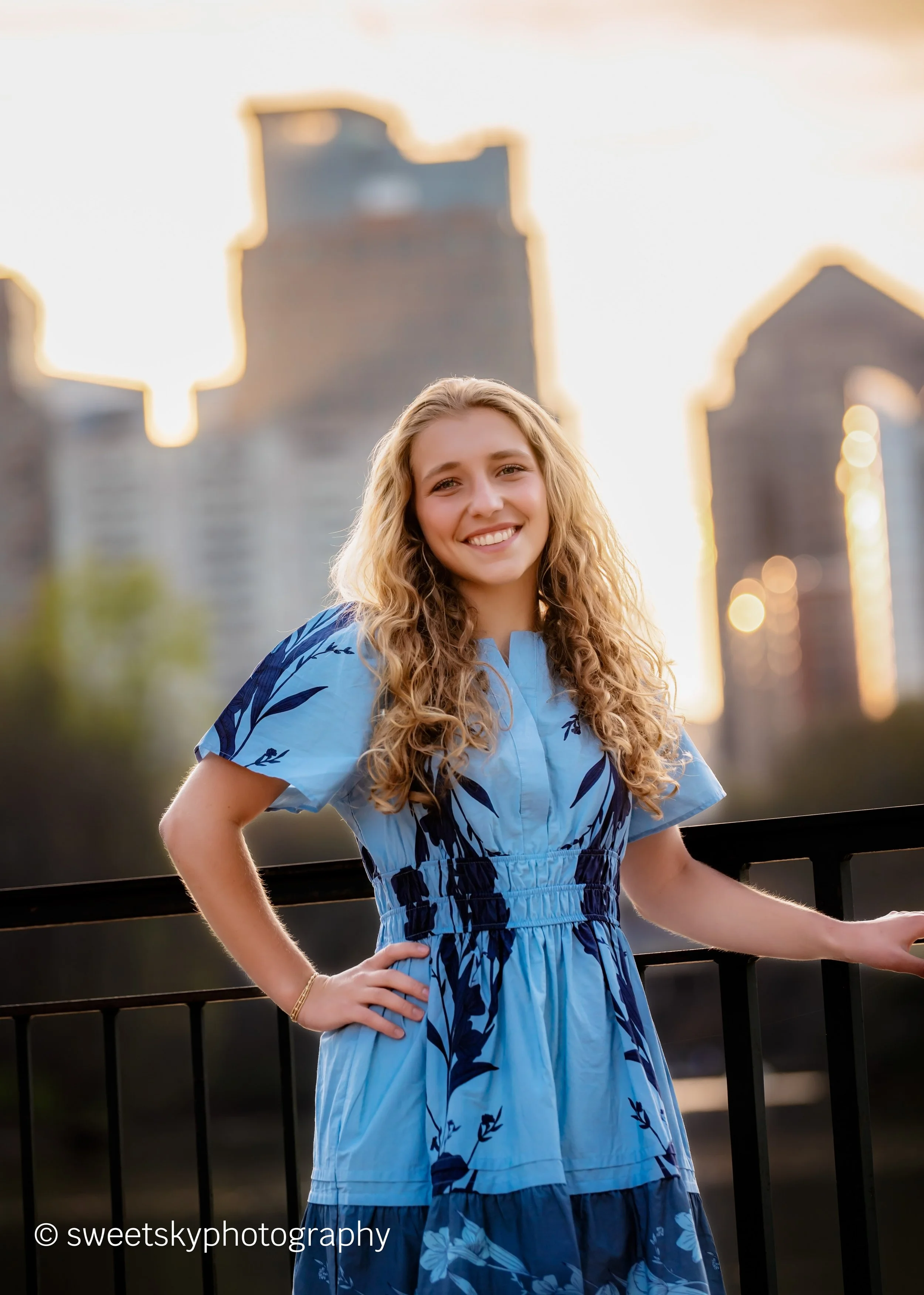A young woman with long curly blonde hair smiling, wearing a blue floral dress, standing outdoors in front of a city skyline at sunset.