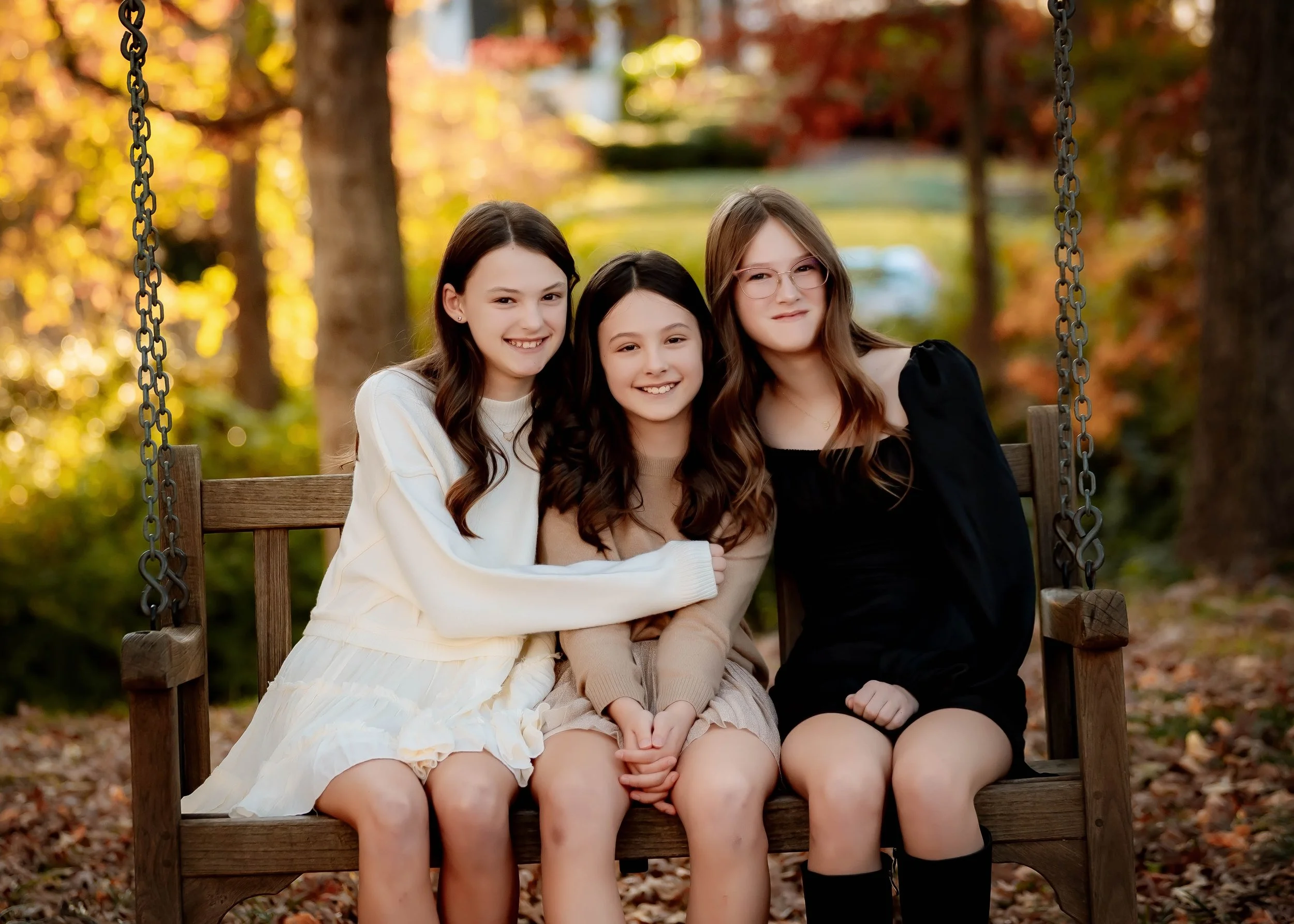 Three young girls sitting on a wooden swing bench outdoors in a park during autumn, smiling at the camera. The background features trees with fall foliage.