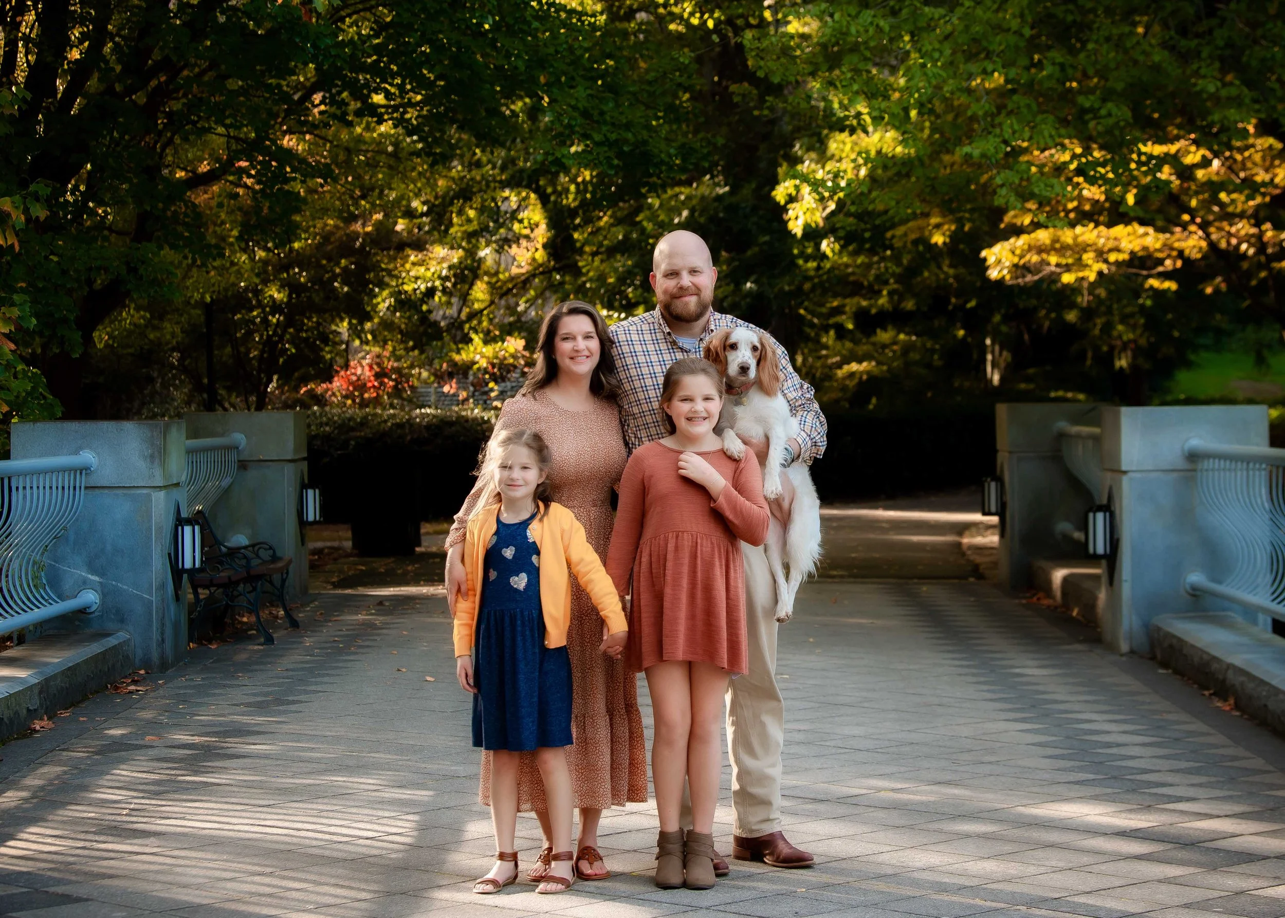 Family of five, including two young girls, an adult woman and man, and a family dog, standing on a pathway in a park with lush green trees and colorful foliage in the background.