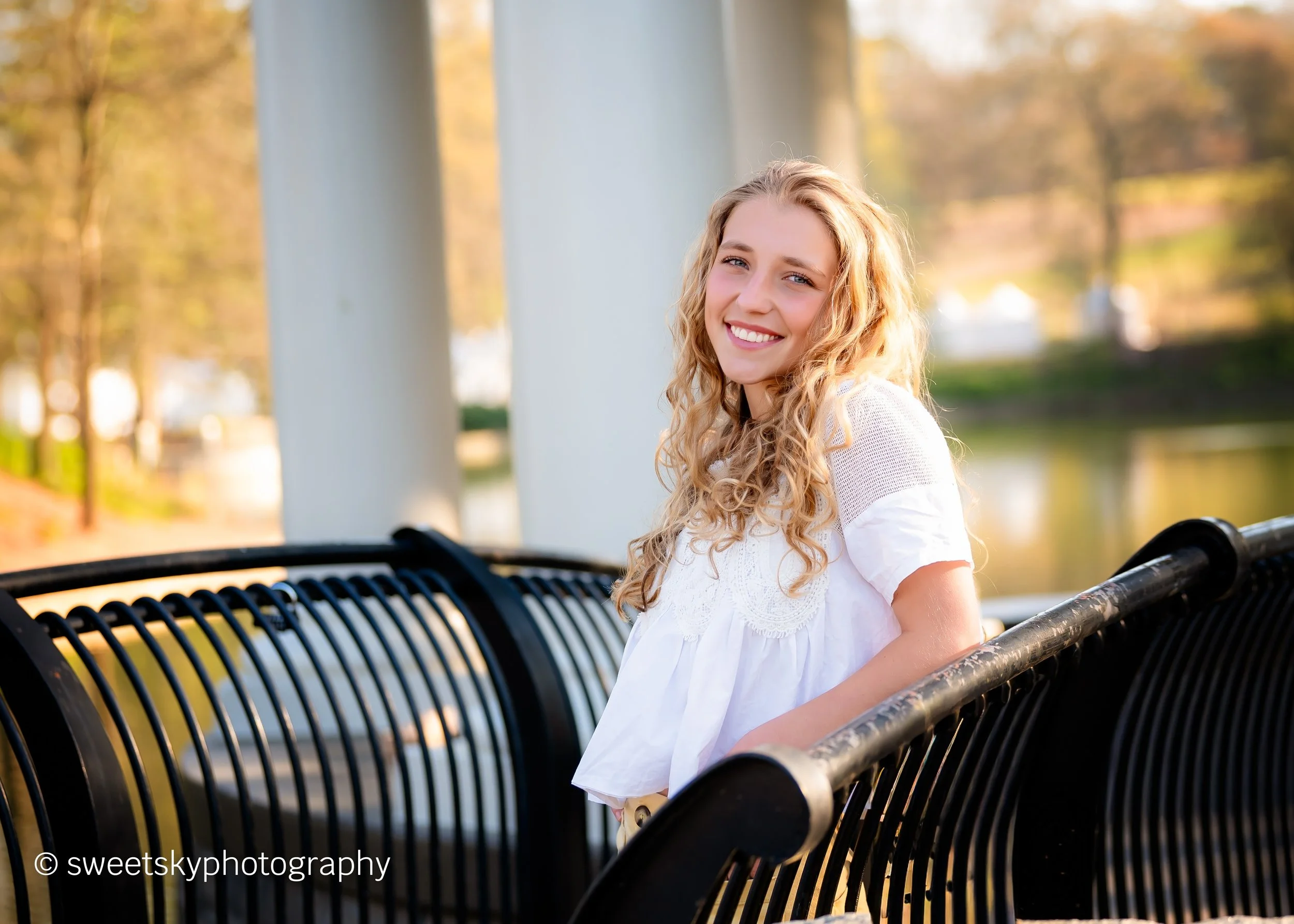 A young woman with curly blonde hair smiling while sitting on a black metal park bench by a river with trees and bridge in the background during daylight.
