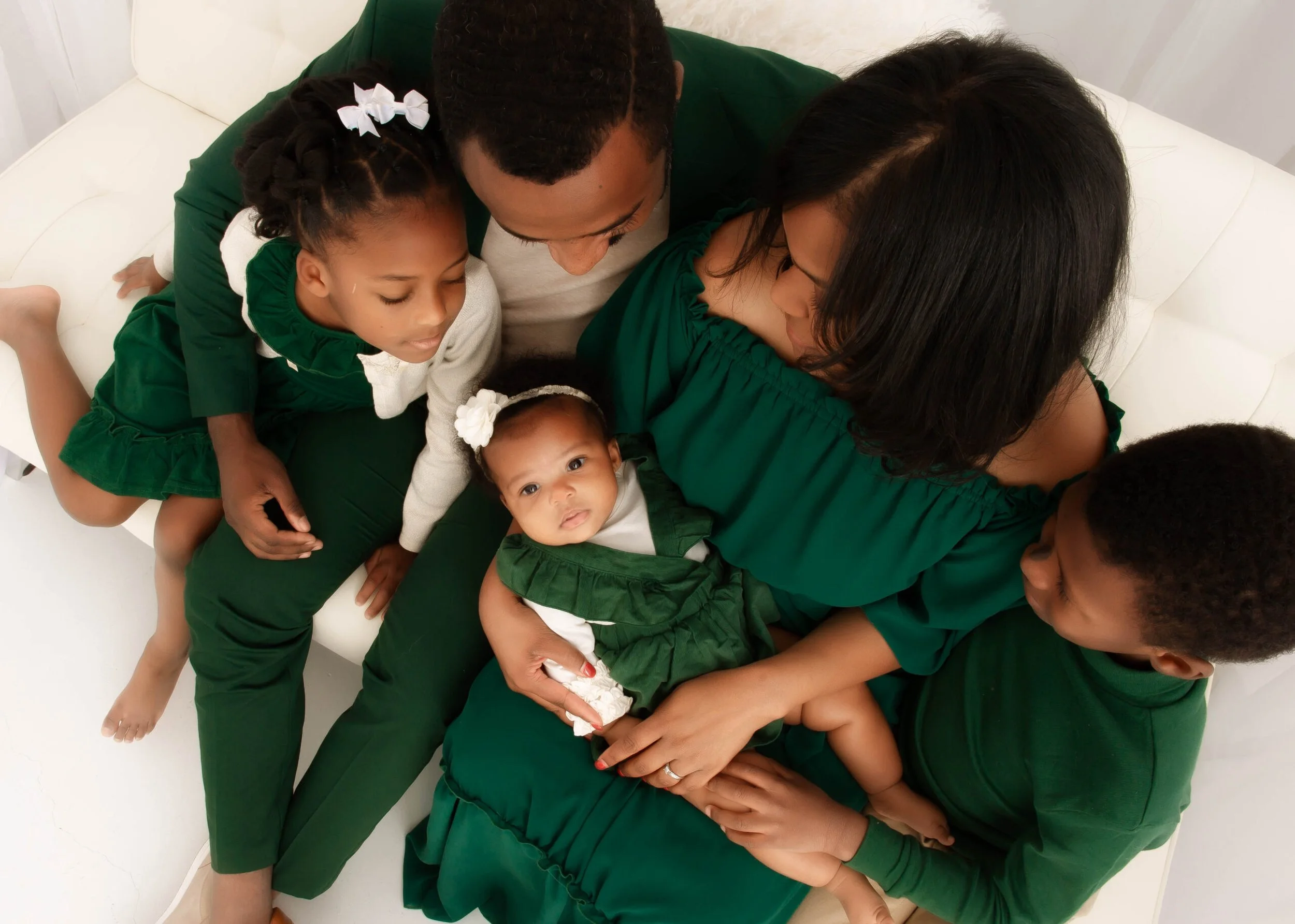 A family of five sitting on a white couch, dressed in green and white, with the mother holding a baby girl, surrounded by children, all looking at the baby.