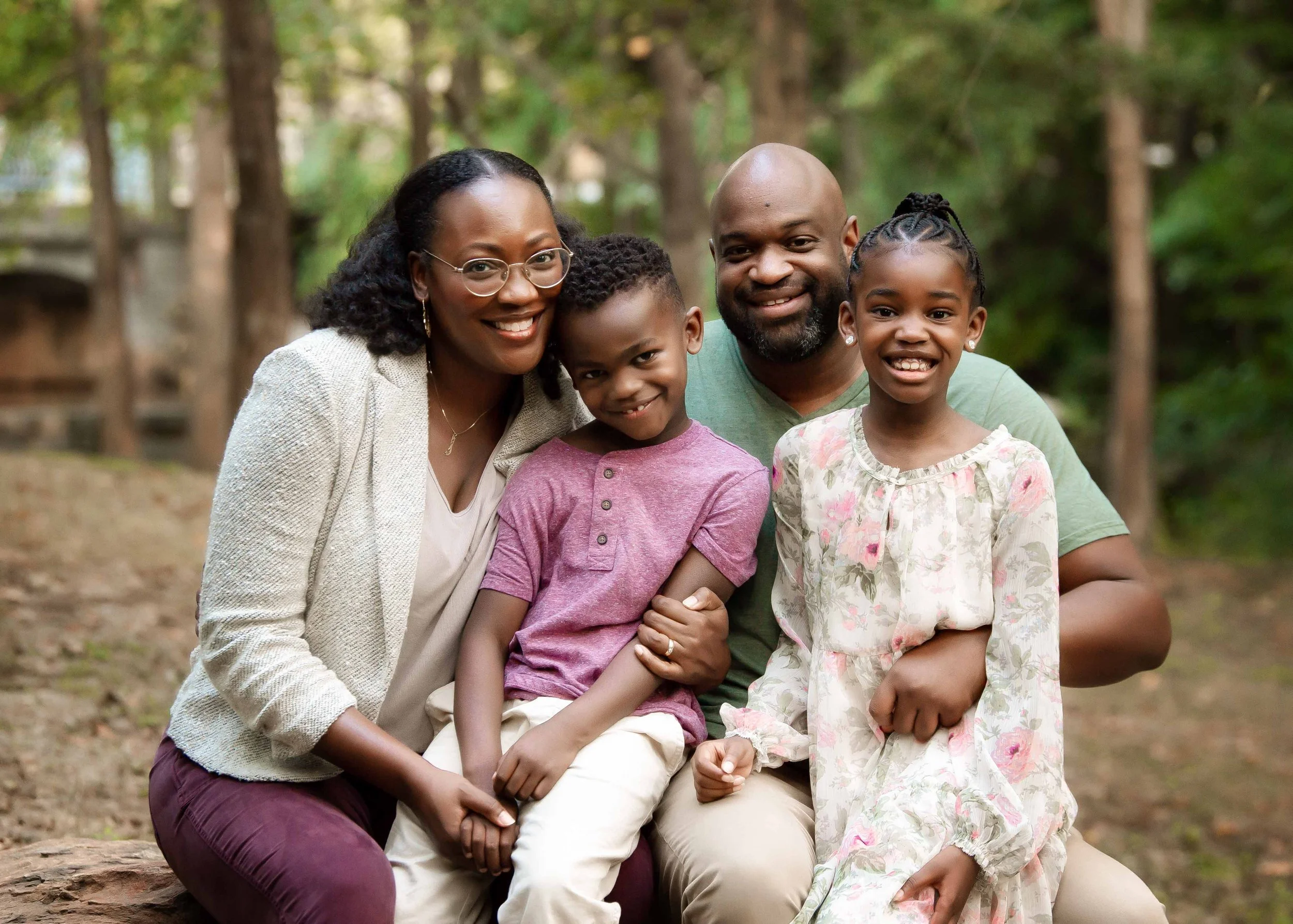 A happy family of four enjoying outdoors in a wooded park, with the parents smiling and two children sitting on their laps, all smiling at the camera.