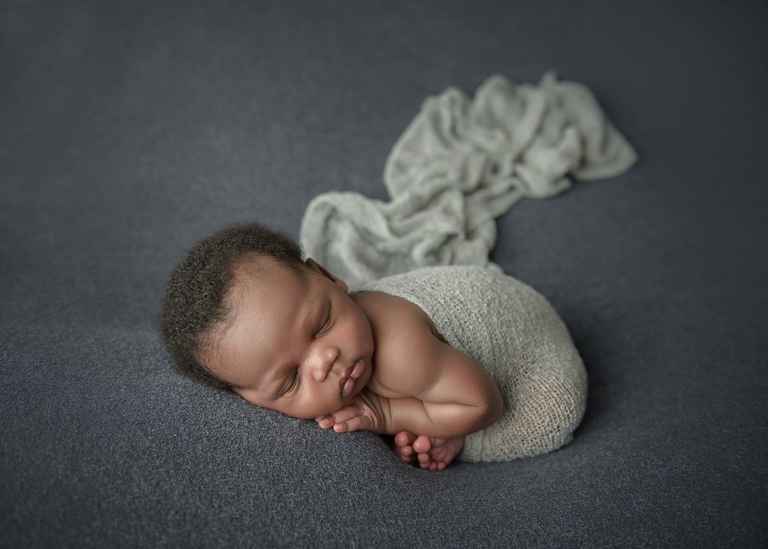 A sleeping baby with brown skin lying on a dark gray surface, wearing a beige knitted outfit, with a soft, light-colored blanket behind him.