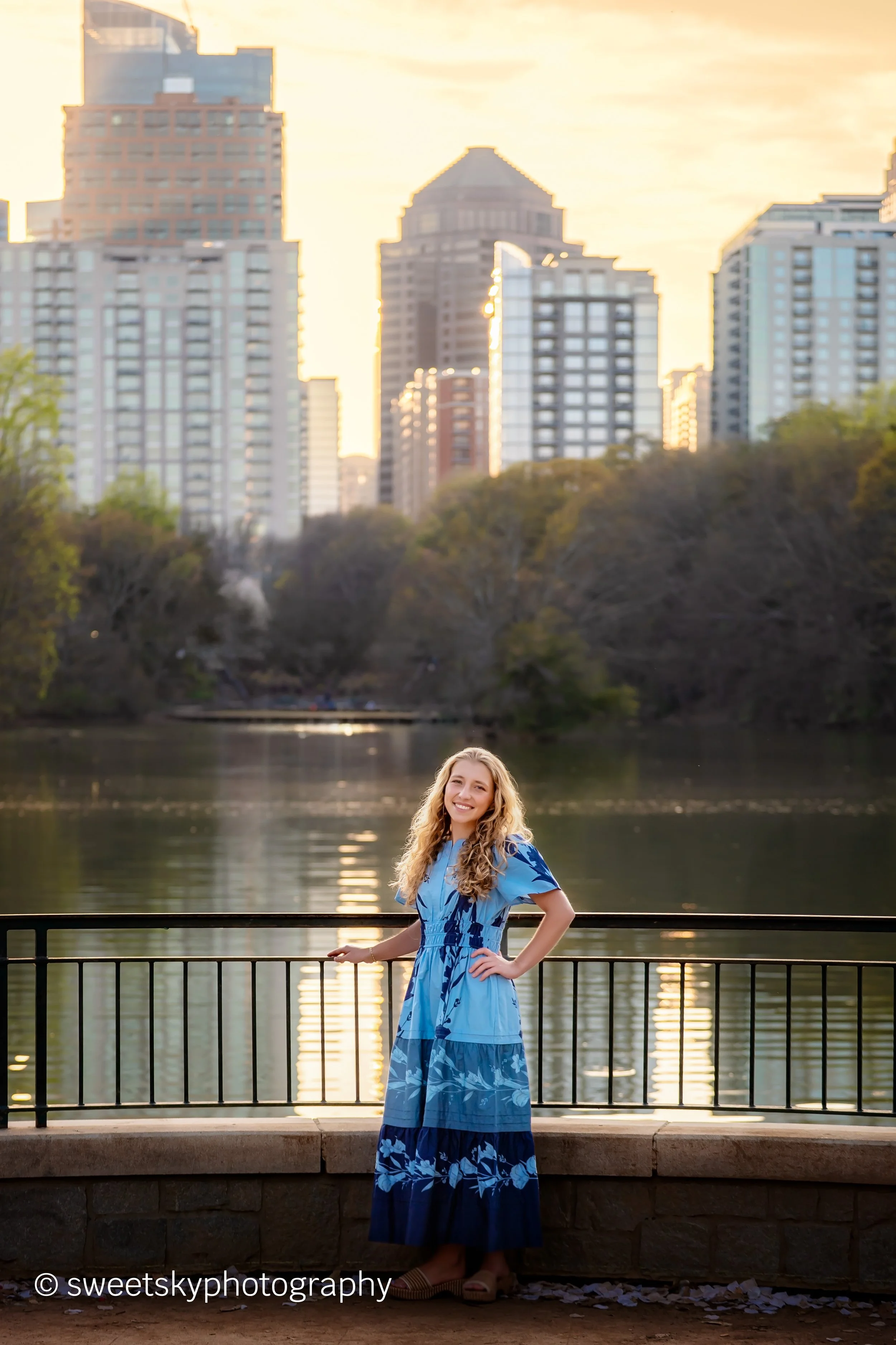A young woman in a blue dress standing by a railing near a river during sunset, with city skyscrapers in the background.