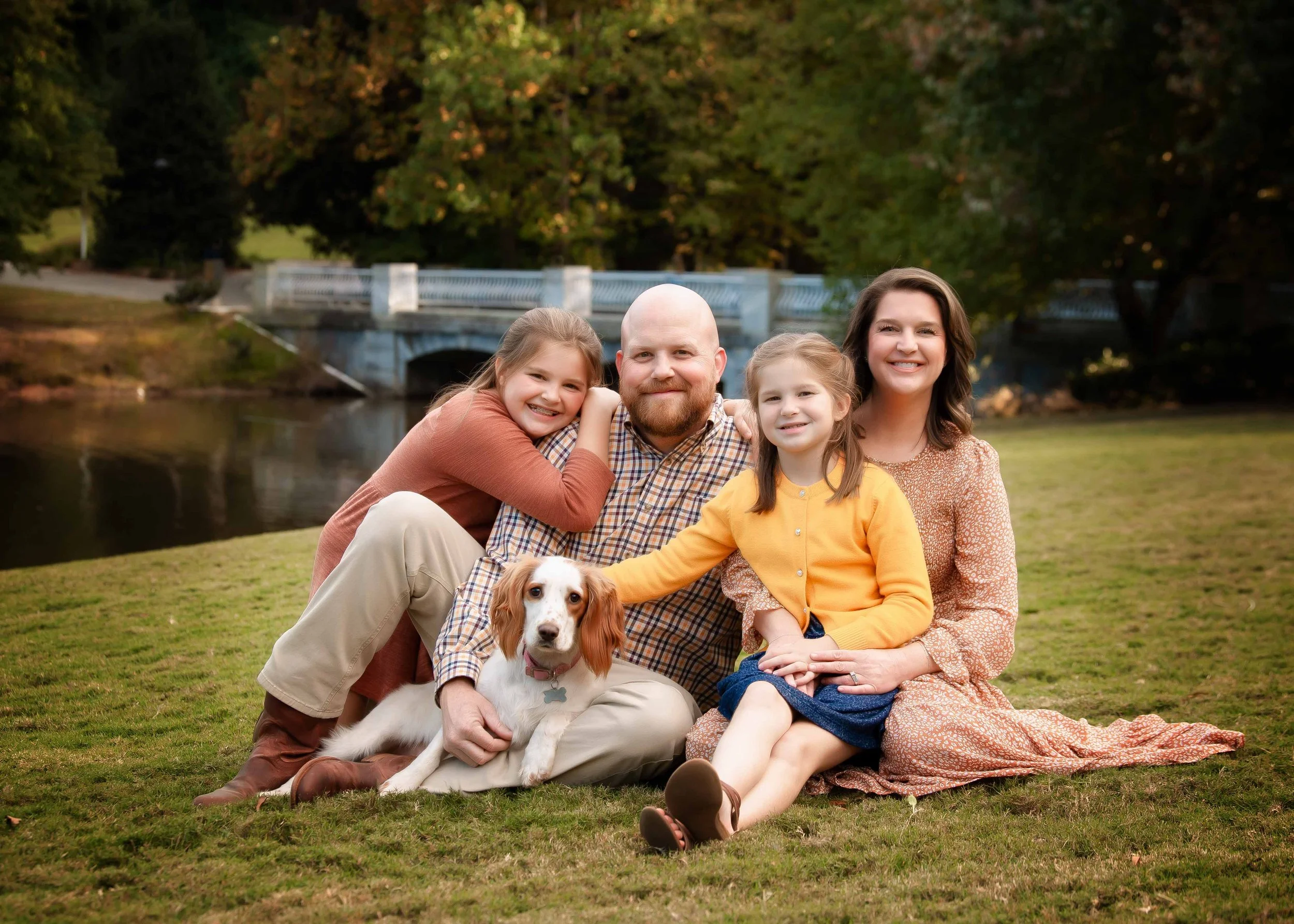 Family of four with two young girls and their dog sitting on grass near a river with trees and a bridge in the background, all smiling.