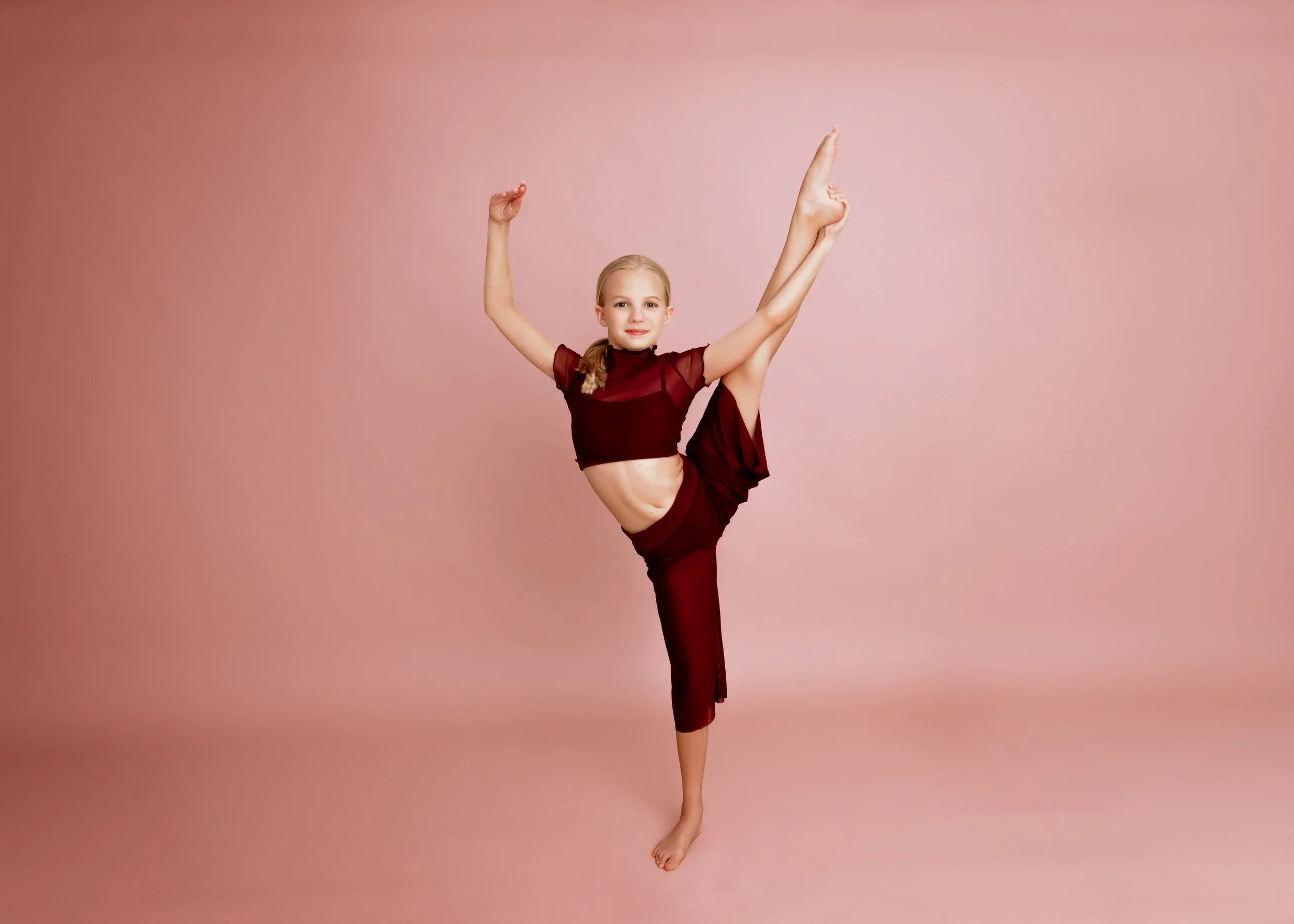 Young girl performing a ballet pose, balancing on one foot with her other leg raised high and arms extended, against a pink background.
