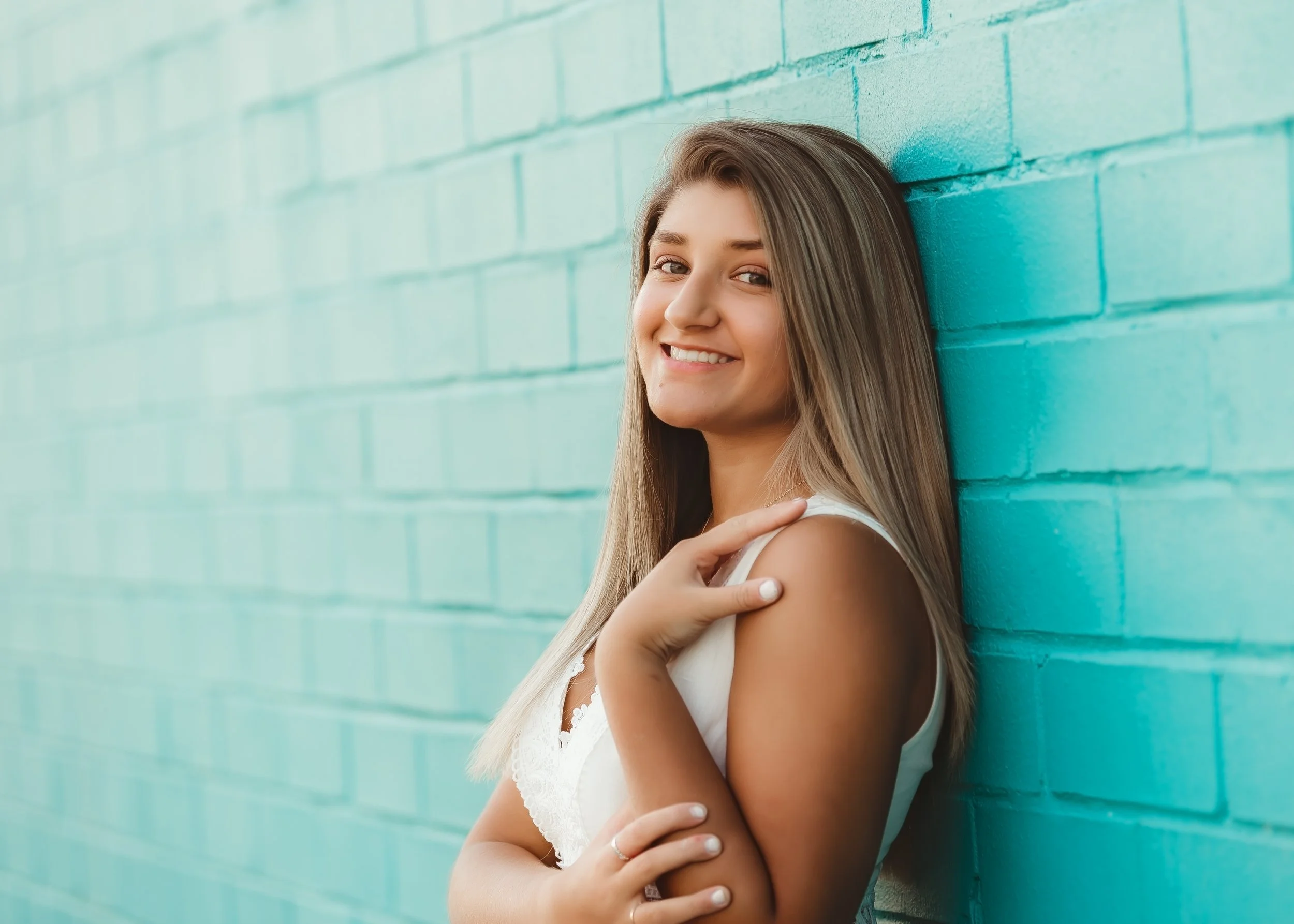 A young woman with long blonde hair smiling and leaning against a turquoise brick wall.