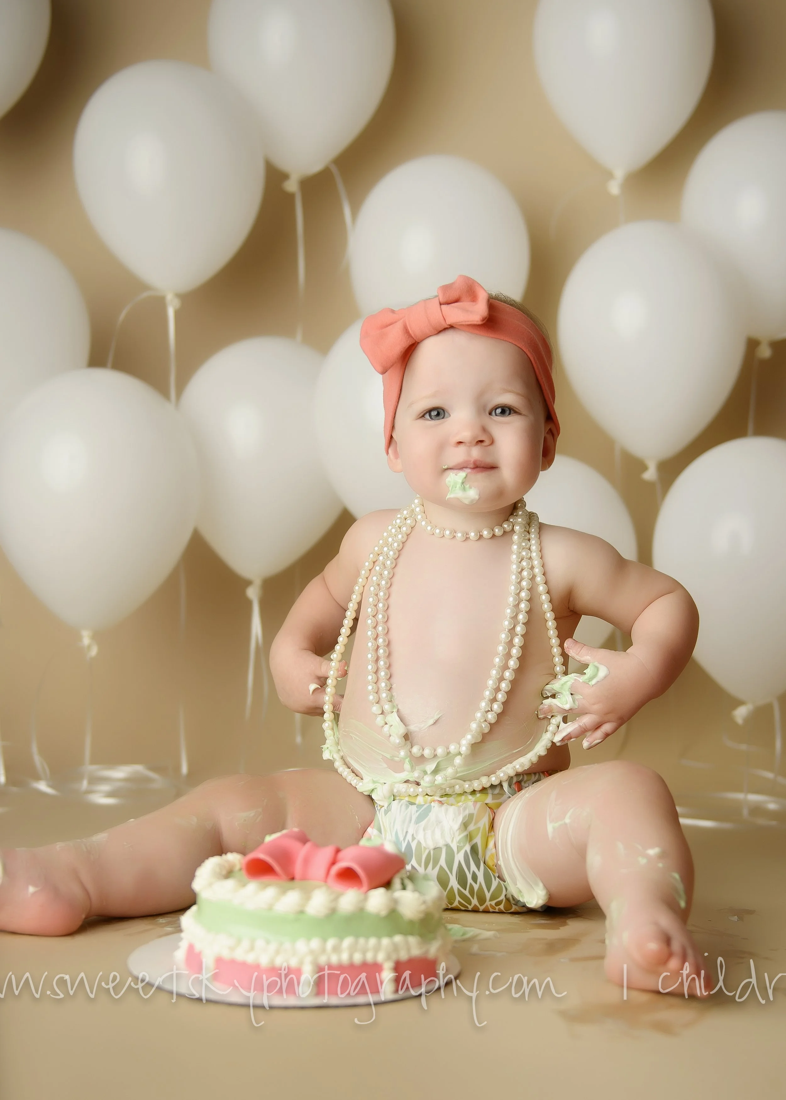A baby sitting on the floor with a small birthday cake in front of her, surrounded by white balloons. The baby is wearing multiple pearl necklaces, a pink headband with a bow, and diaper, with cake crumbs and frosting on her face, hands, and clothes.