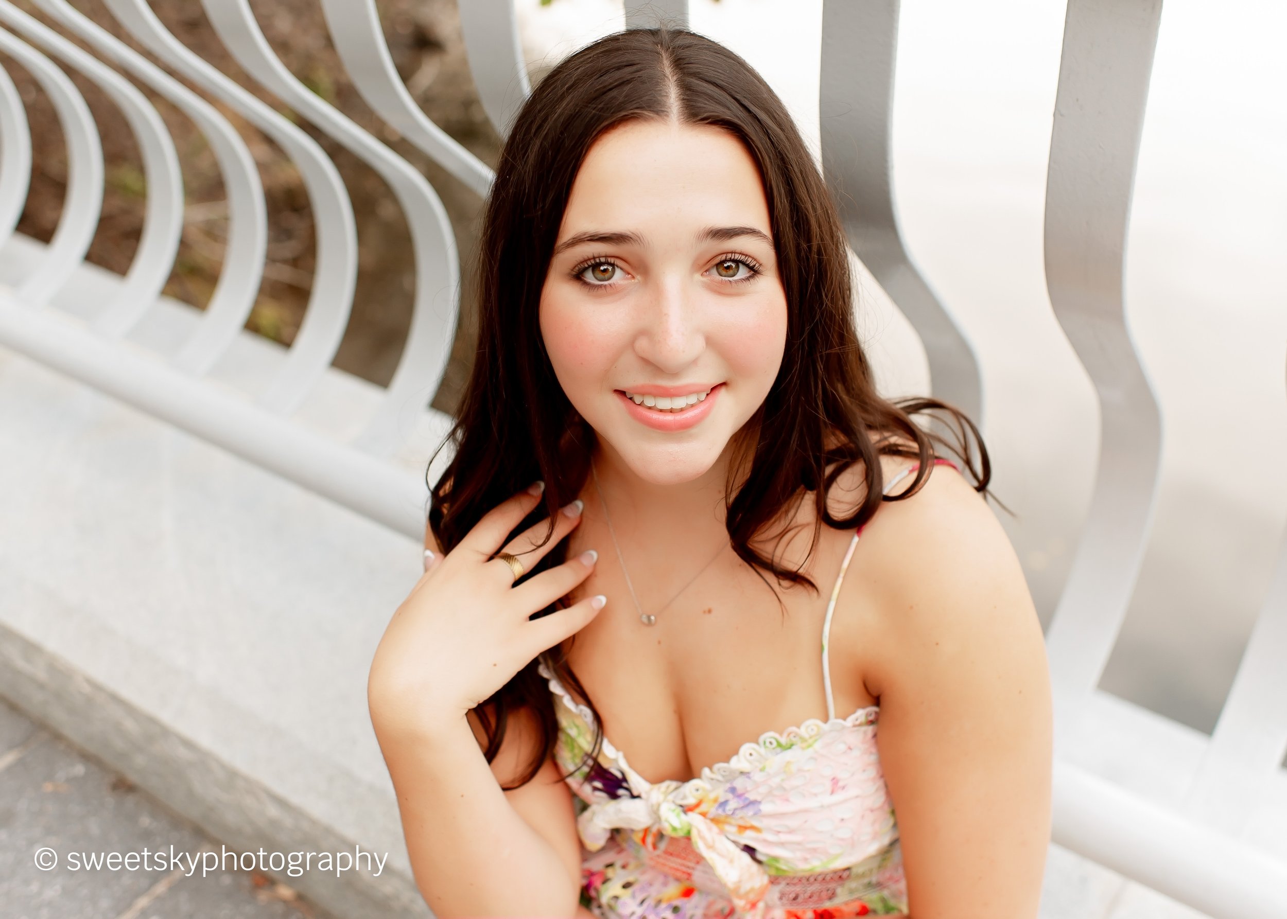 A young woman with long brown hair and light skin sitting on a concrete surface near a white curved metal fence, smiling and touching her hair with her right hand, wearing a floral dress and a necklace.