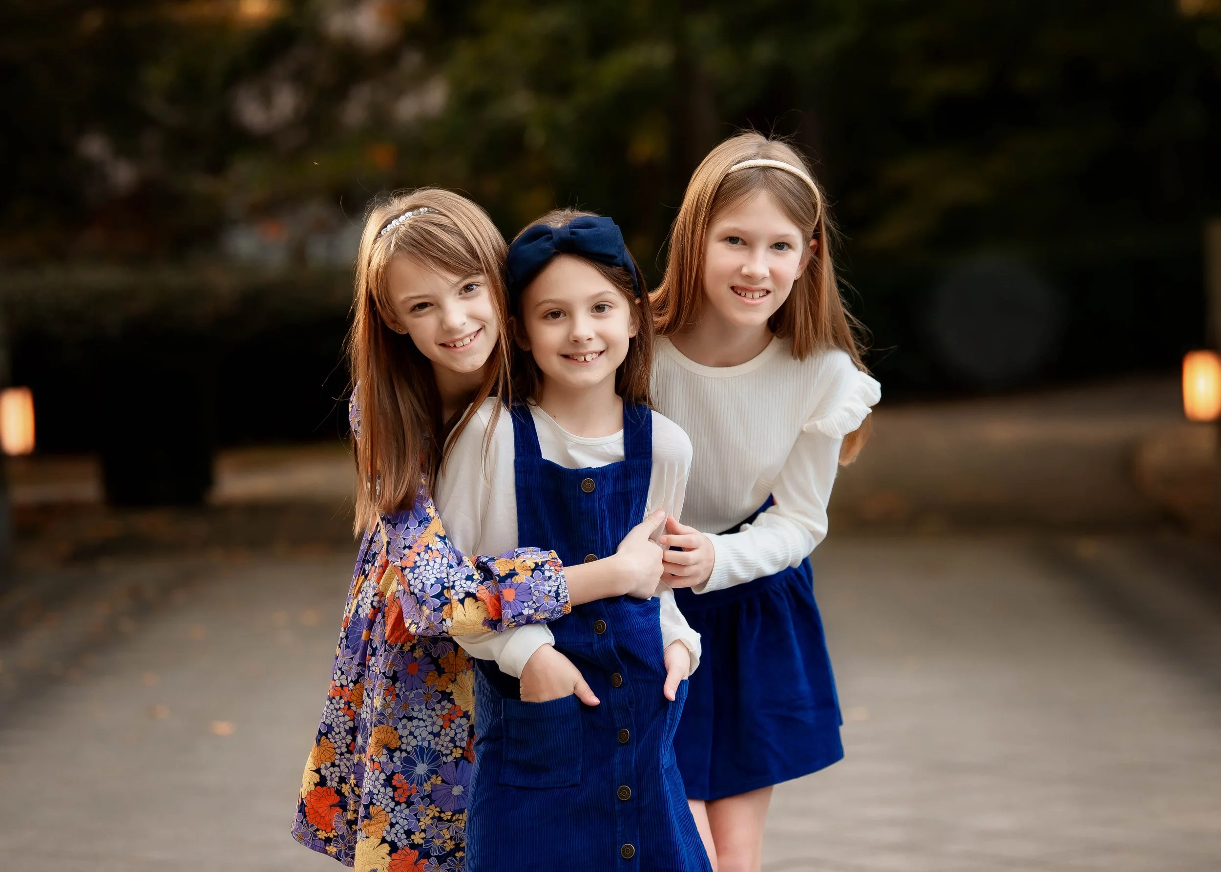 Three young girls standing outside in a park during evening, smiling at the camera.