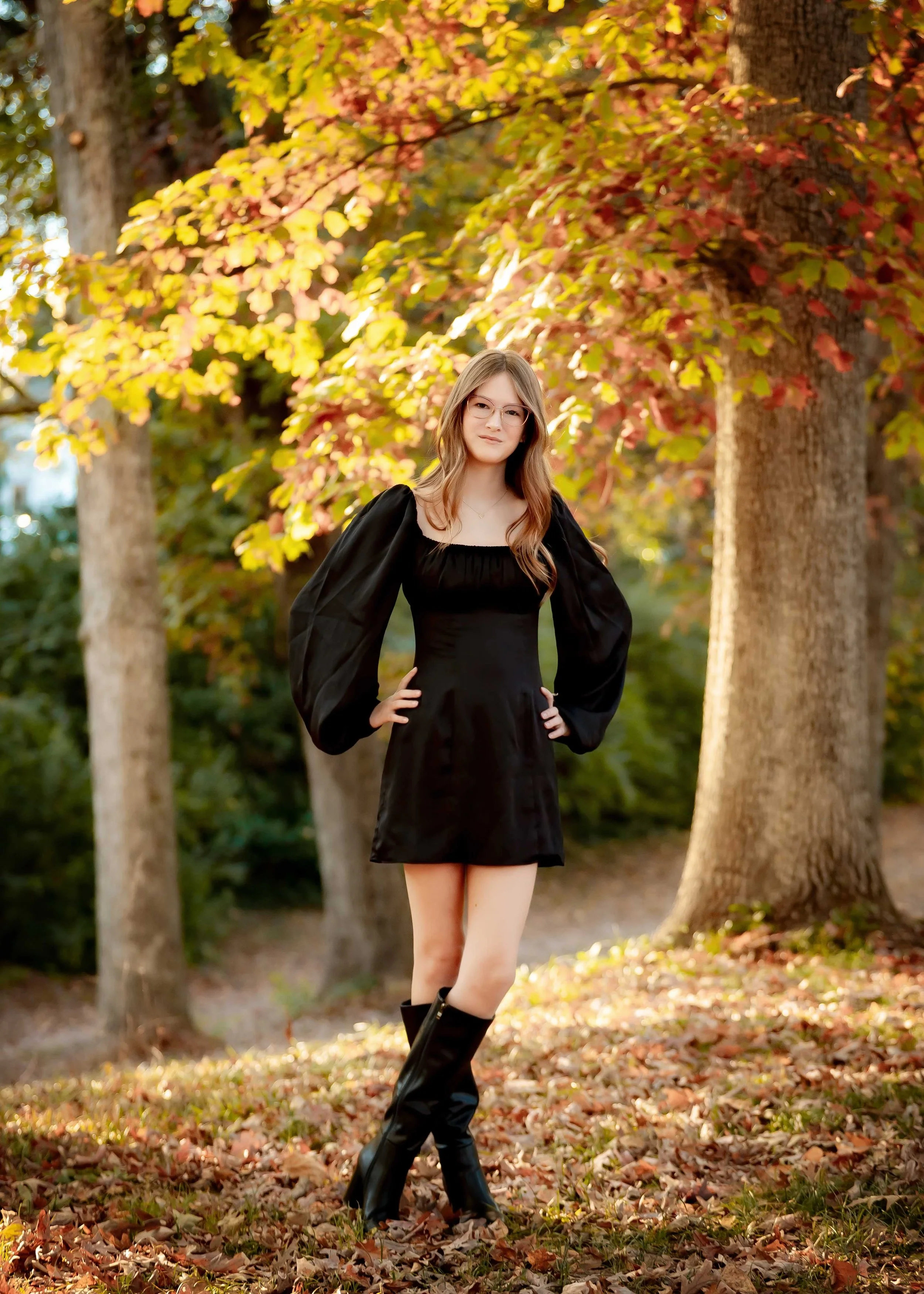 A young woman with long hair, glasses, and a confident pose standing outdoors among trees with autumn leaves, wearing a black dress and knee-high boots.