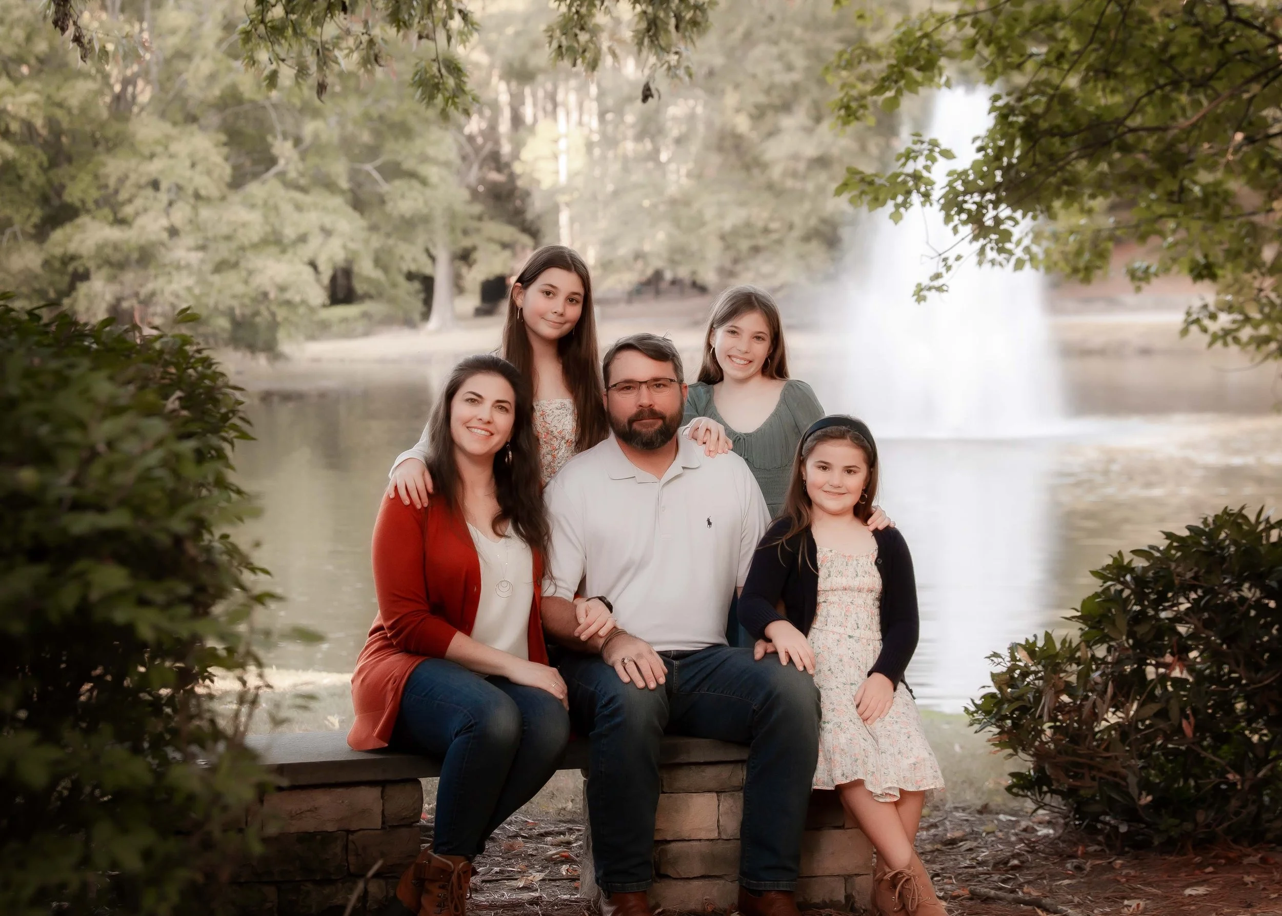 Family portrait of five members, two adult women, one adult man, and two young girls, sitting and standing outdoors near a lake with a waterfall in the background and surrounded by trees.