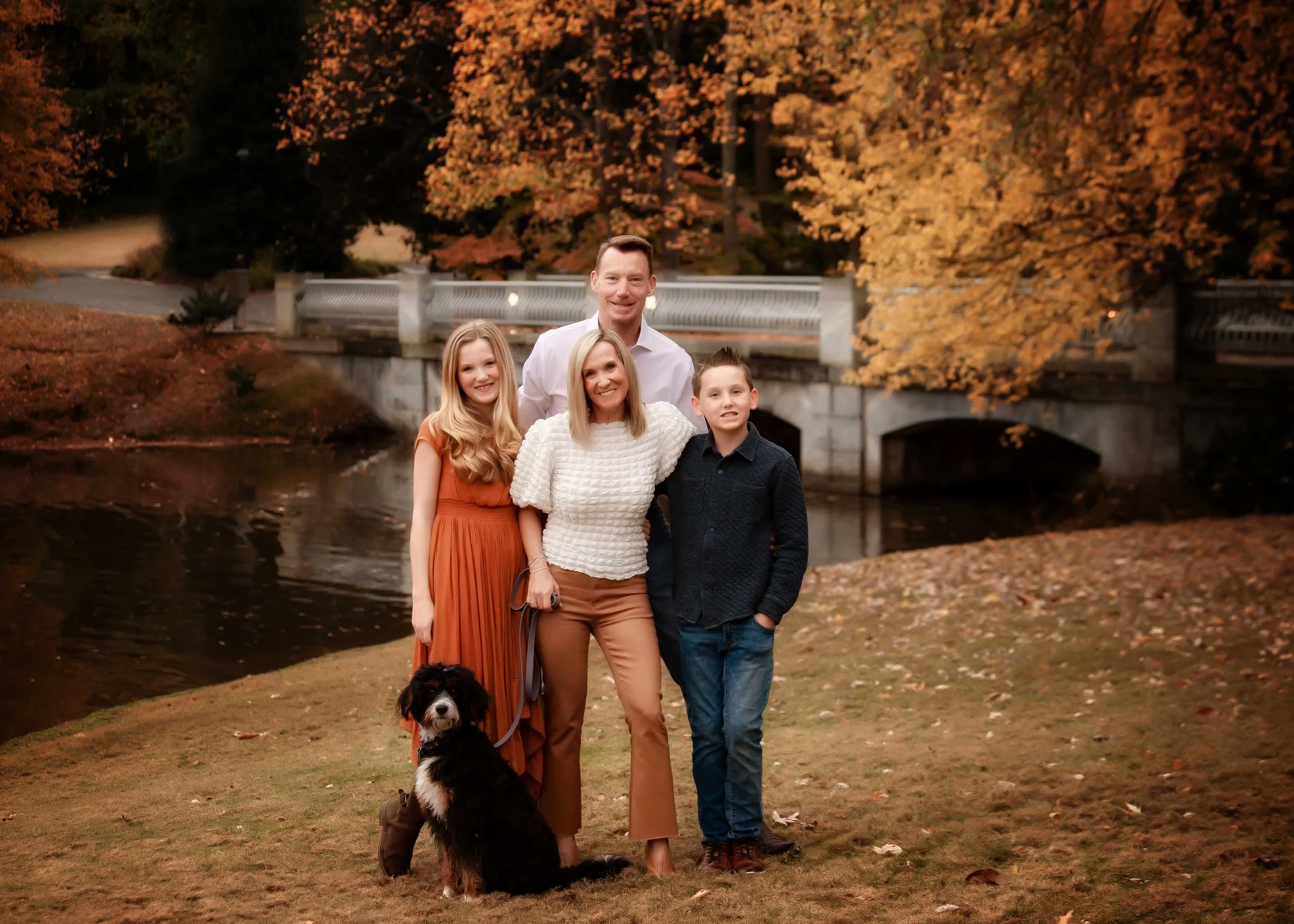A family of four with a dog standing outdoors near a river, with autumn trees and a bridge in the background.