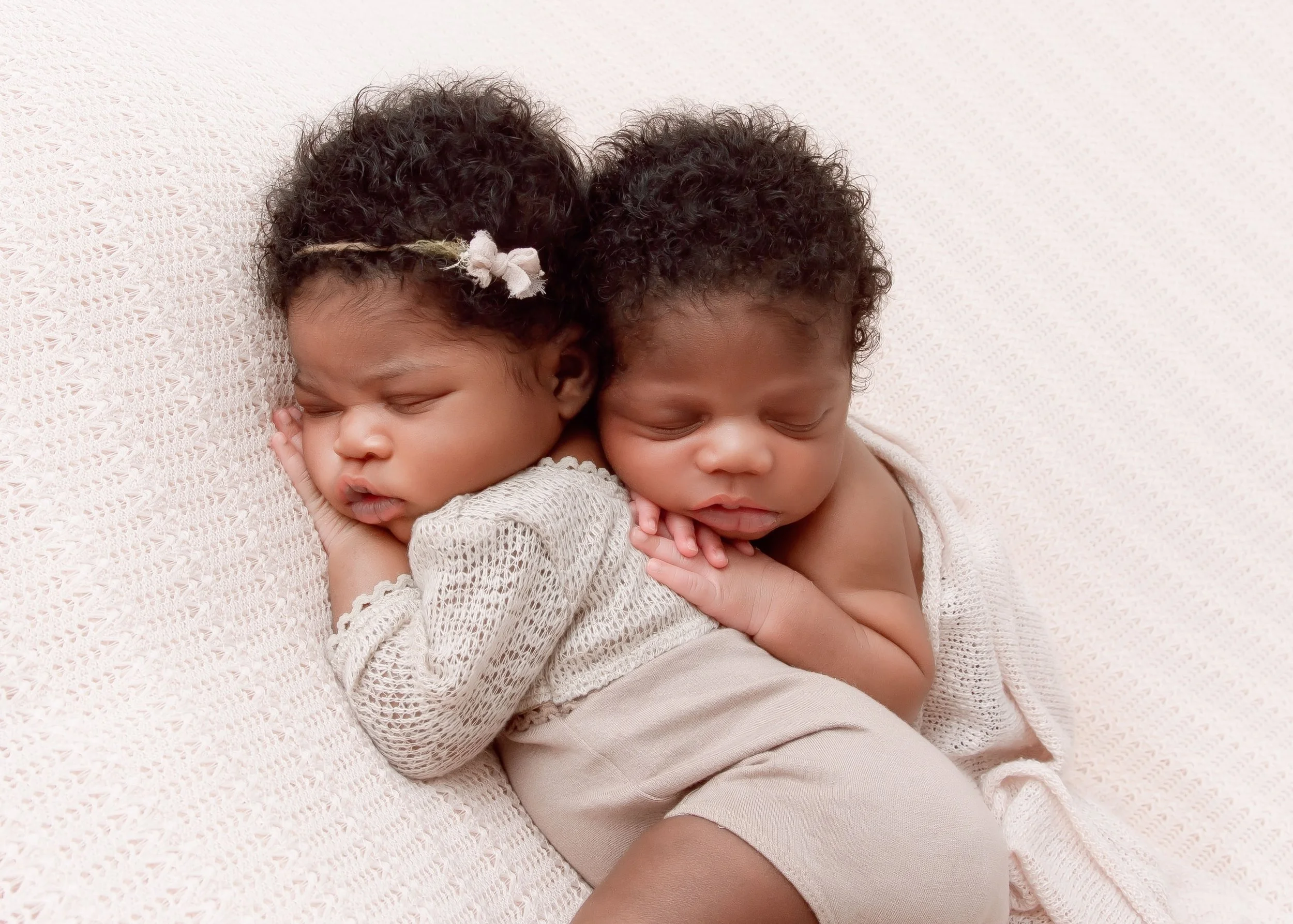 Two young children, a girl and a boy, are cuddling and sleeping together on a soft, light-colored blanket. They are resting peacefully, with the girl wearing a headband with a flower and the boy with curly hair.