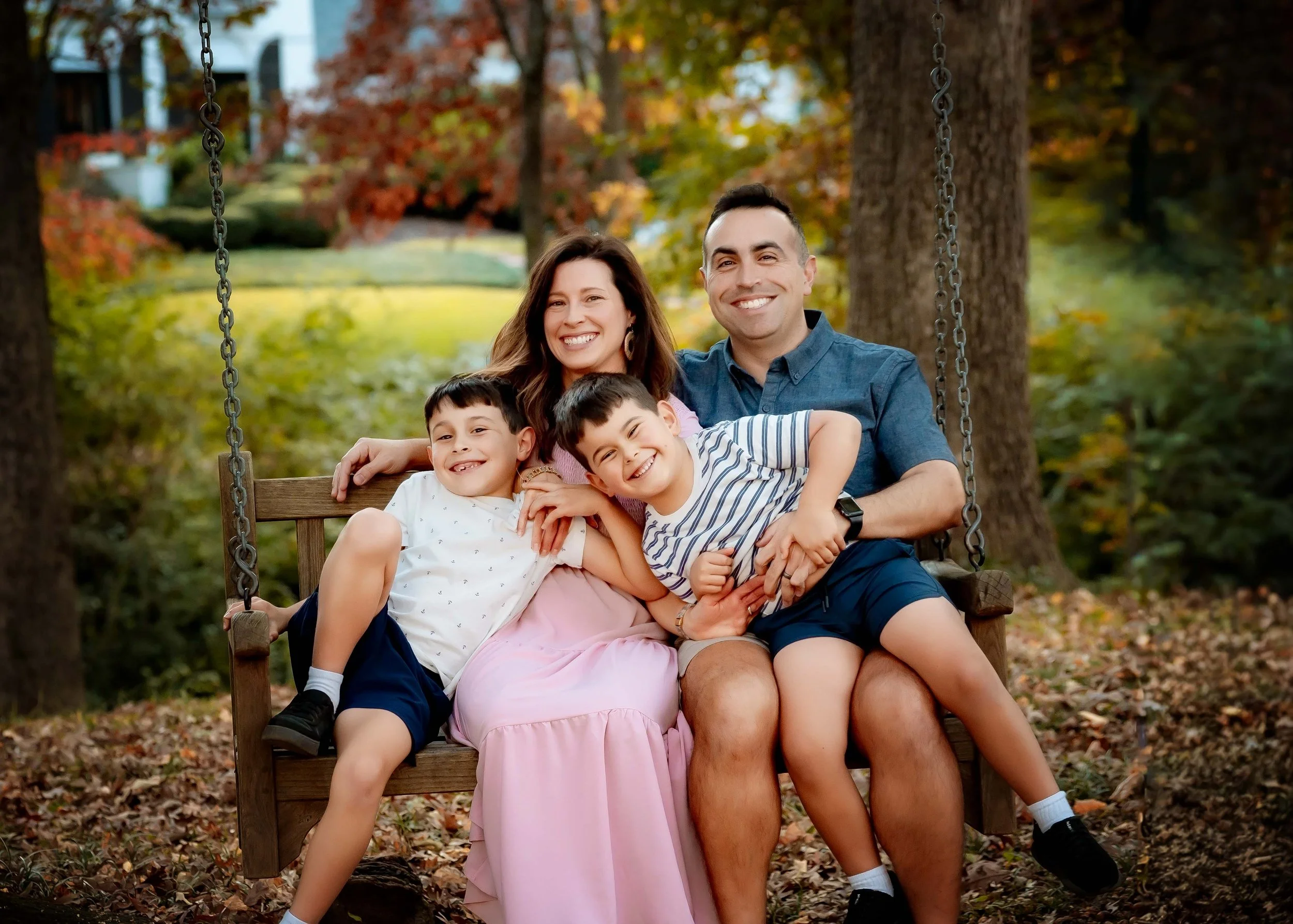 A happy family of four sitting on a wooden swing in a park during fall, with colorful autumn trees in the background. The mother and father are smiling, with the sons sitting in their laps.