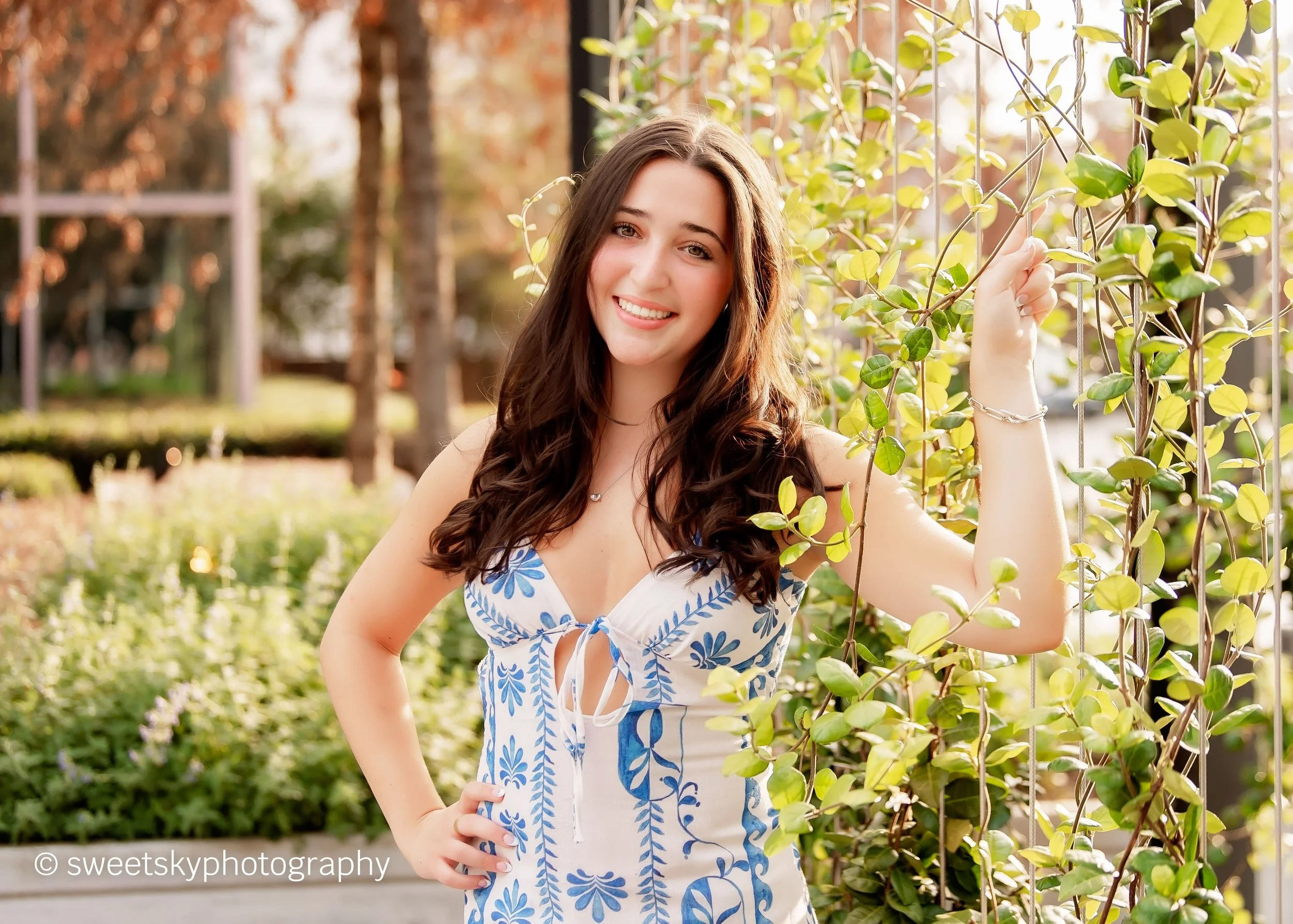 A young woman with long, wavy brown hair smiling and posing outdoors beside a vine-covered structure in a park or garden during daytime.