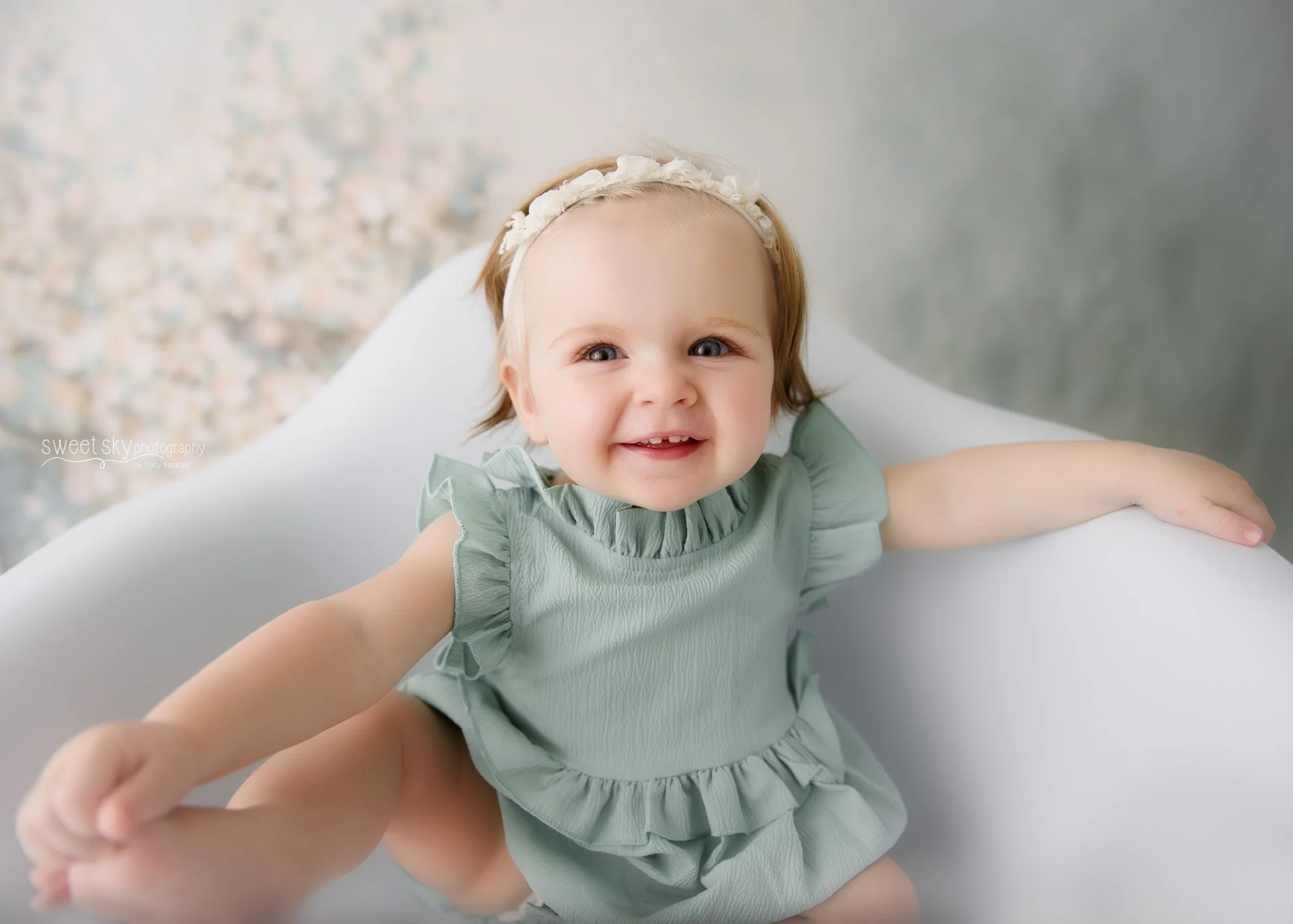A young girl with light hair, blue eyes, wearing a green dress and a white floral headband, smiling and reaching out with her arm.