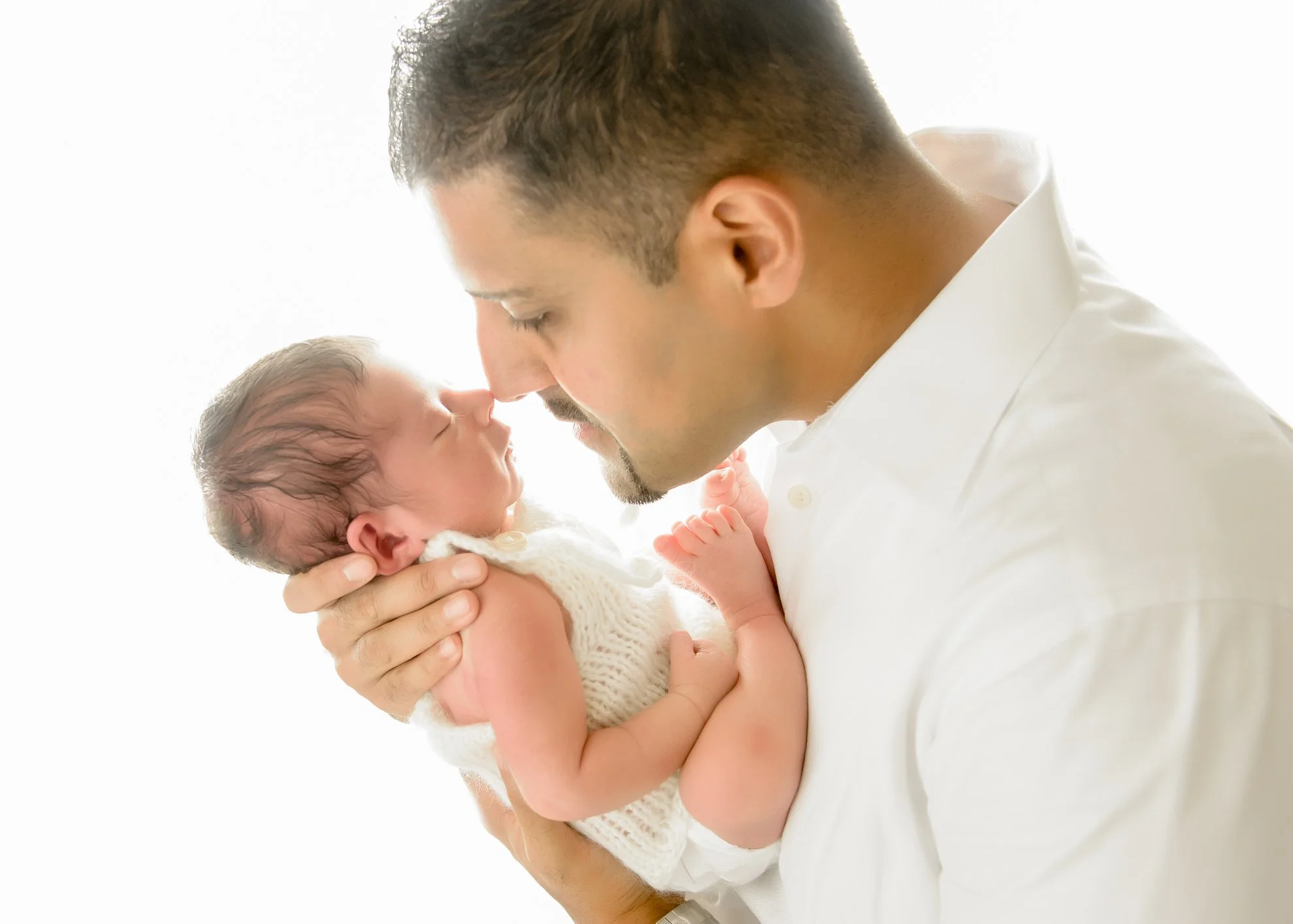 A man holding a newborn baby close to his face, with their foreheads touching, both with their eyes closed, in a bright setting.
