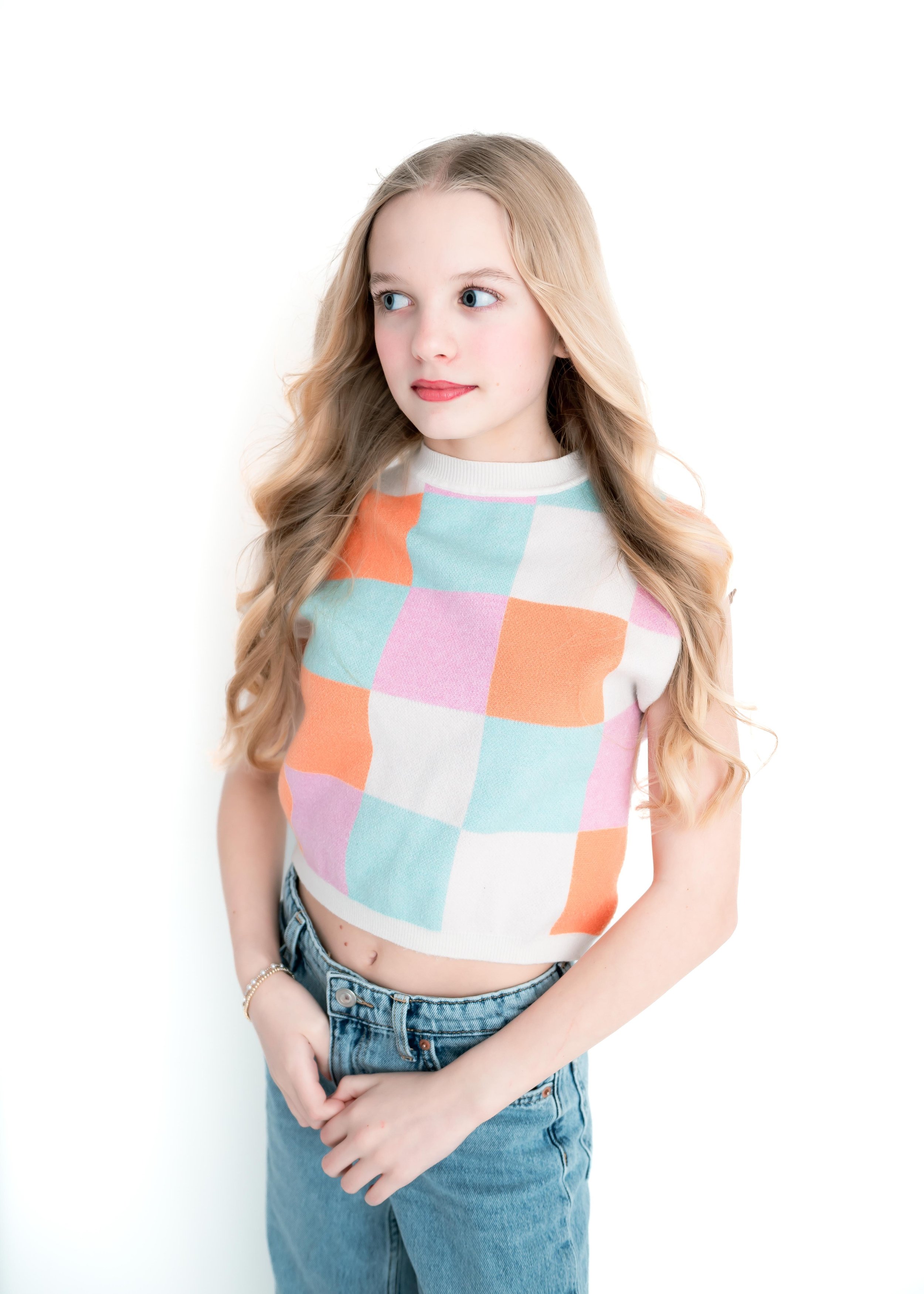 A young woman with long blonde hair and blue eyes wearing a sleeveless pastel-colored checkered sweater and high-waisted jeans.