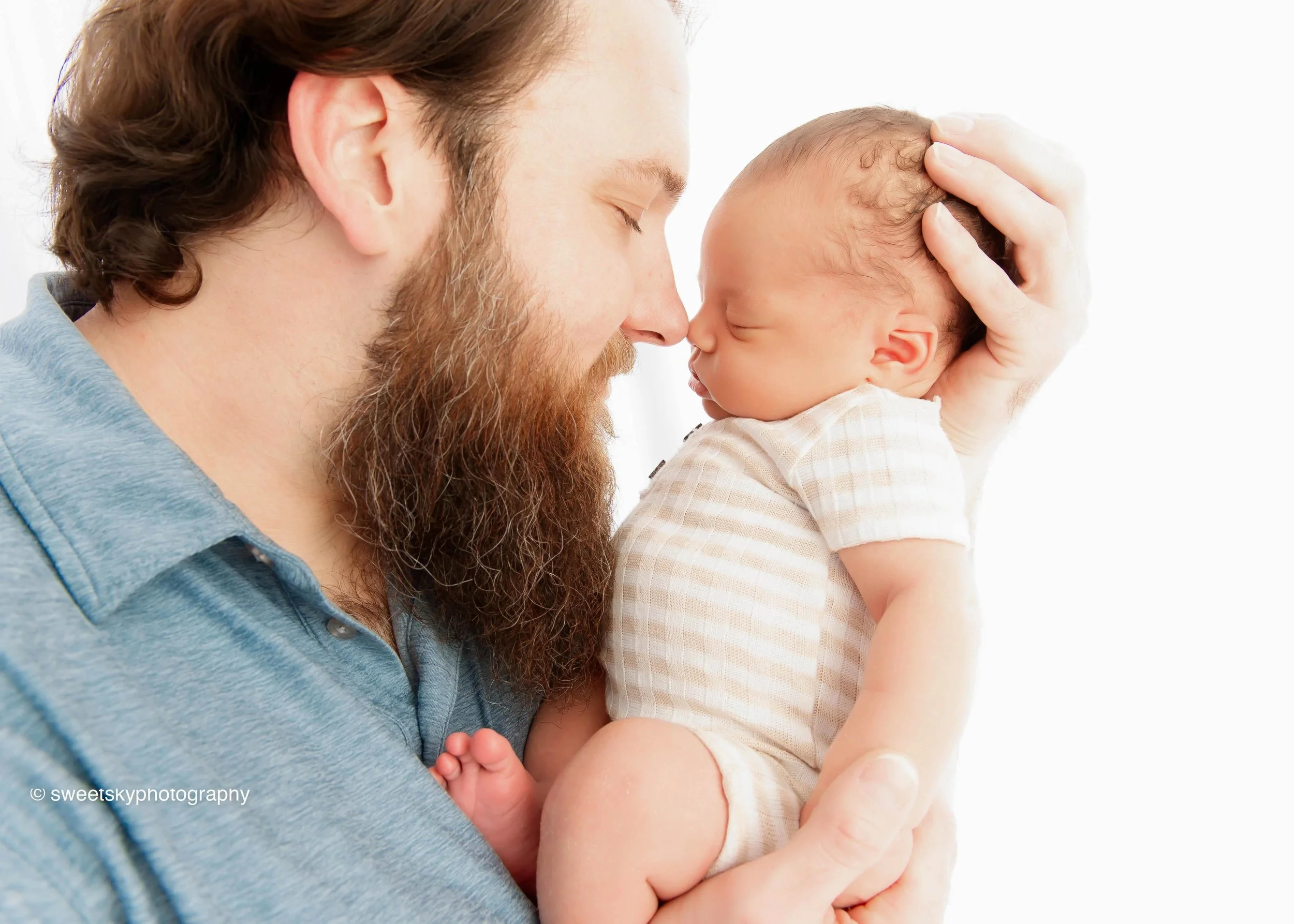 A man with a beard holds a newborn baby close to his face, both with noses touching, in a tender moment. The baby is wearing a light-colored striped outfit.