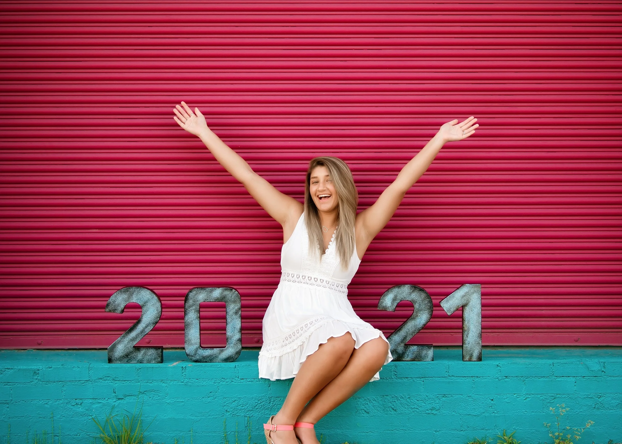 Young woman sitting in front of a pink shutter with the numbers 2021, wearing a white dress, smiling, with arms raised.