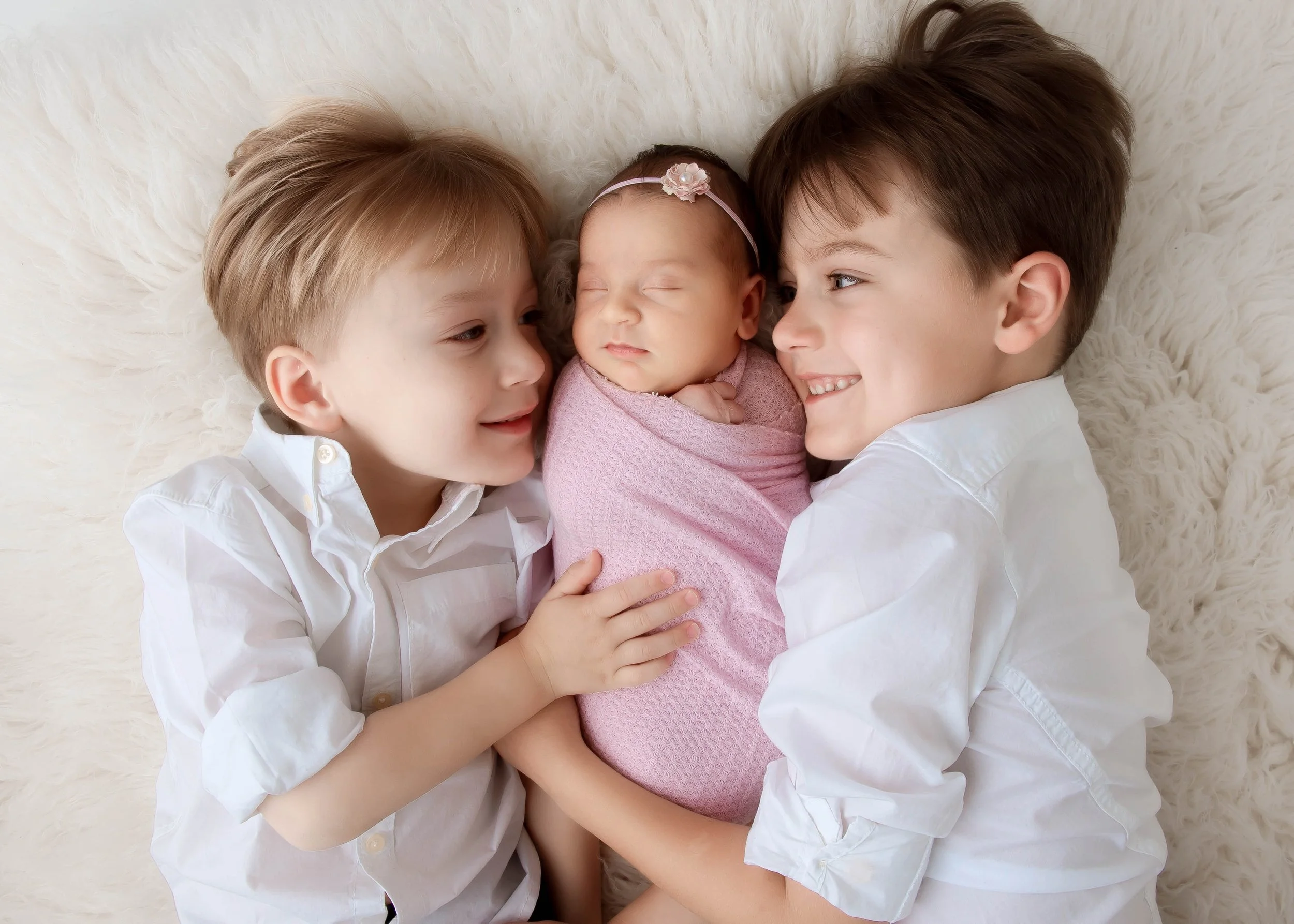 Two young boys and a newborn girl lying together on a soft surface, with the boys smiling and the girl sleeping.