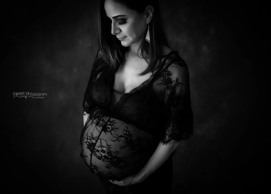 A pregnant woman wearing a black lace top, looking down with a gentle smile, against a dark background.