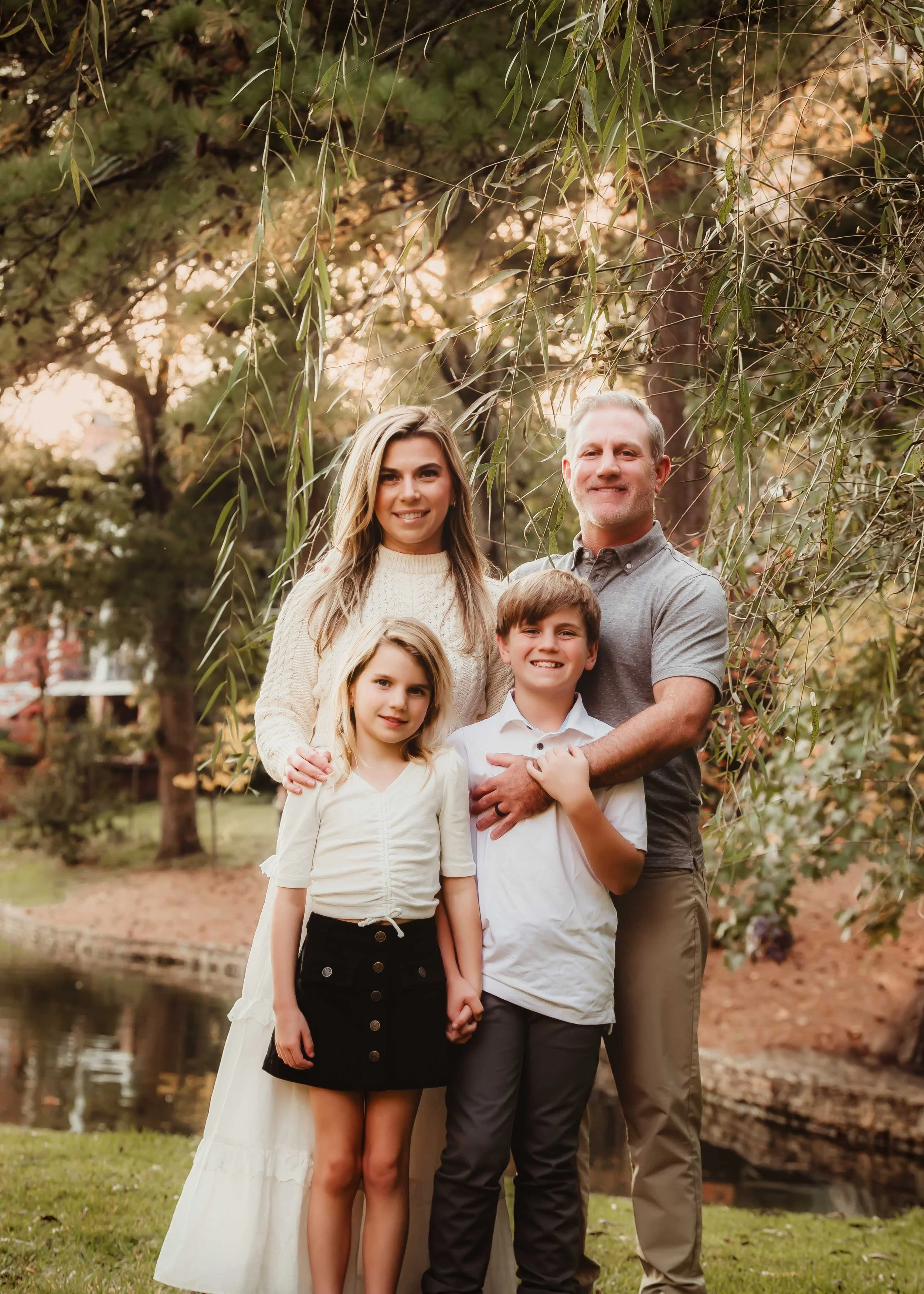 A family of five posing outdoors near a pond with trees in the background, smiling at the camera.