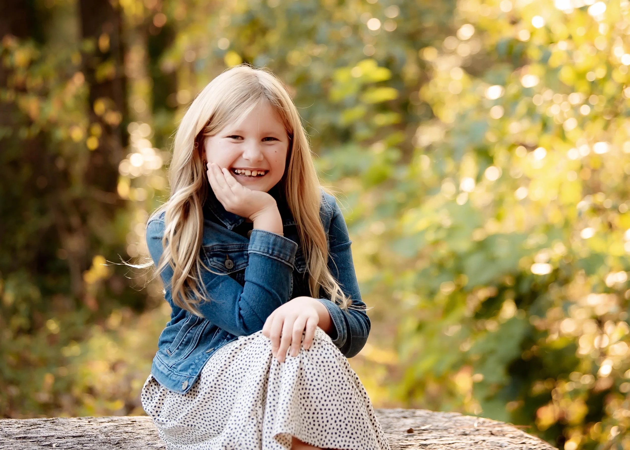 A young girl sitting on a log outdoors, smiling, wearing a denim jacket and a polka dot skirt, with a background of green trees and sunlight.