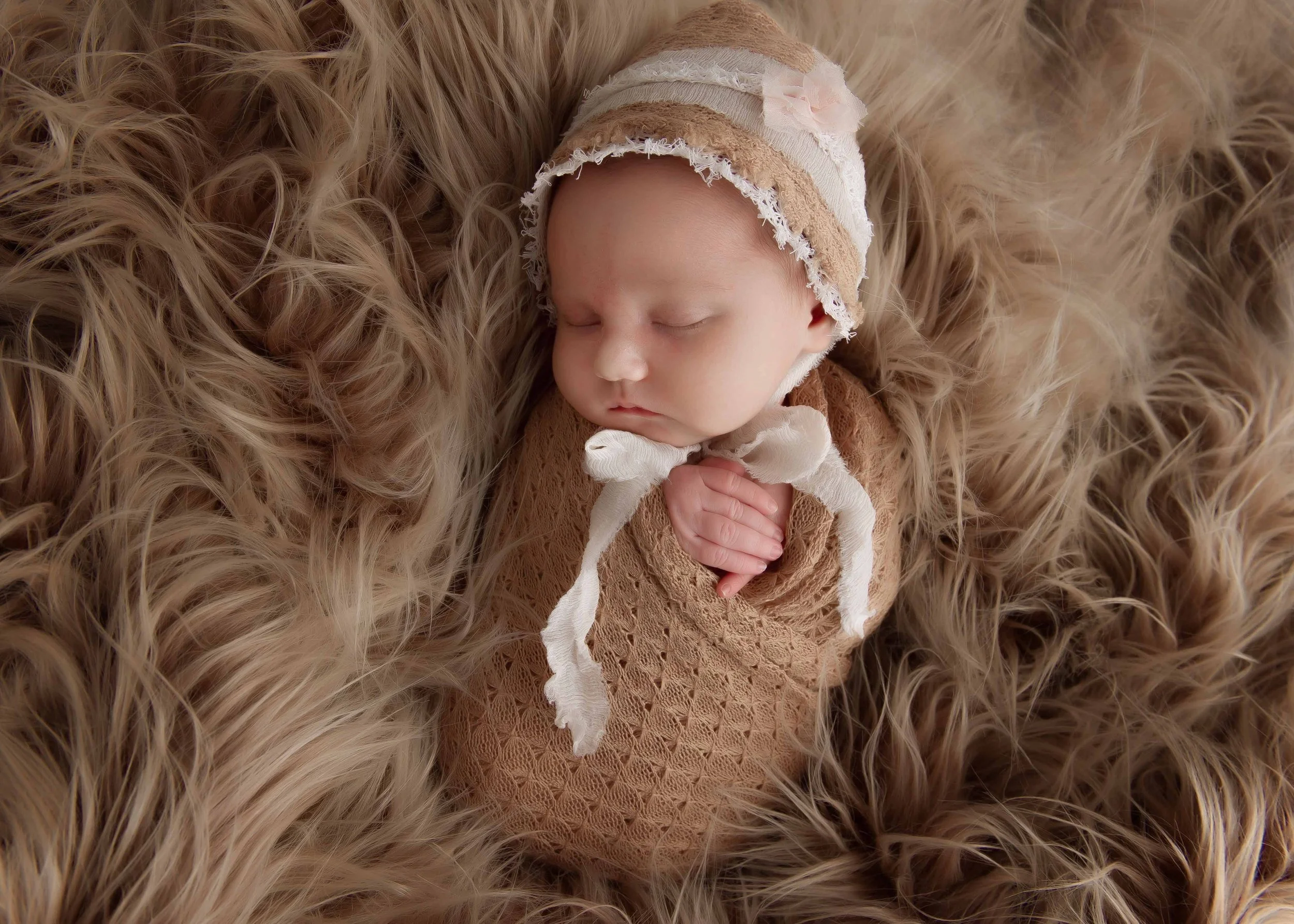 Close-up of a newborn baby sleeping on a fluffy, textured, light brown blanket, wearing a matching beige lace outfit and a striped hat with pink flowers.