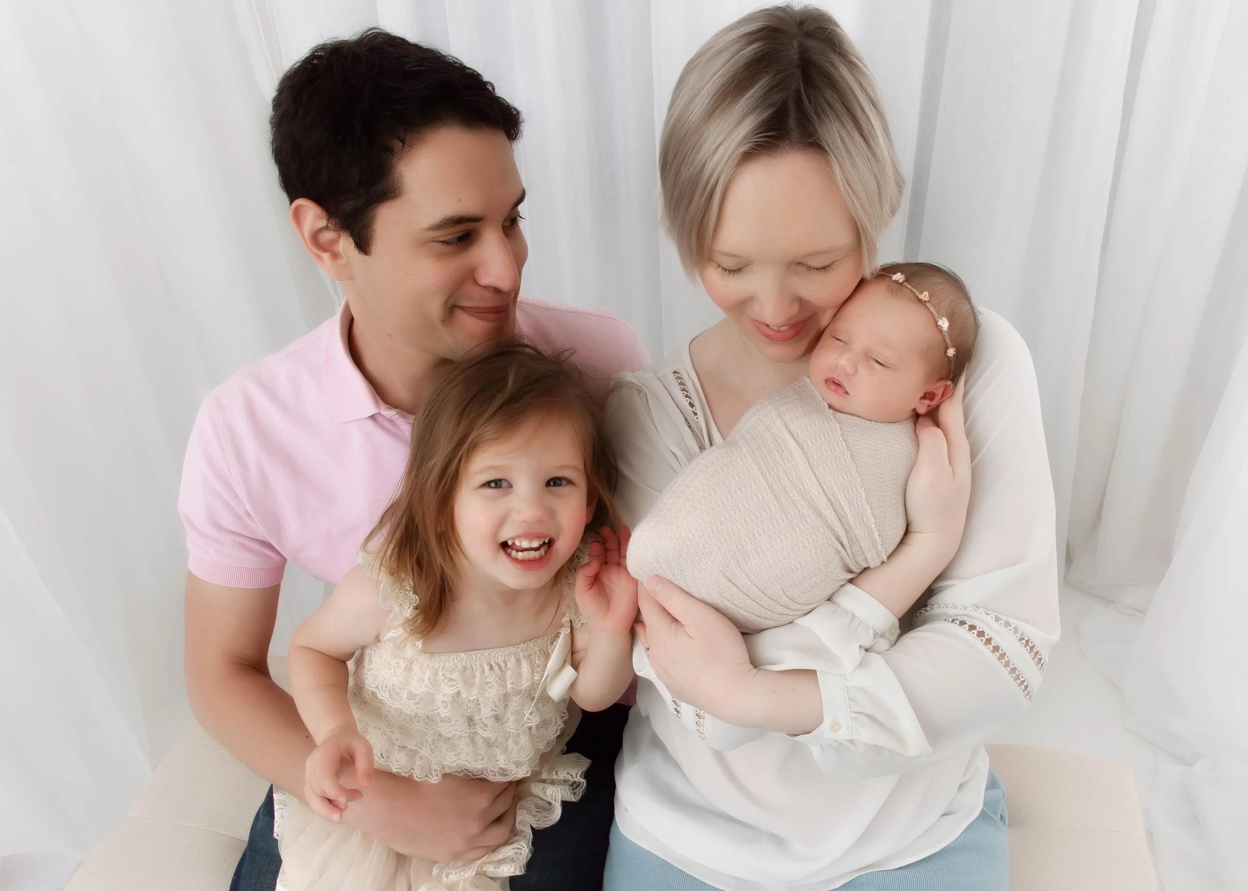 Family of four, including mother, father, young daughter, and newborn baby, smiling and cuddling together in a bright, white room.