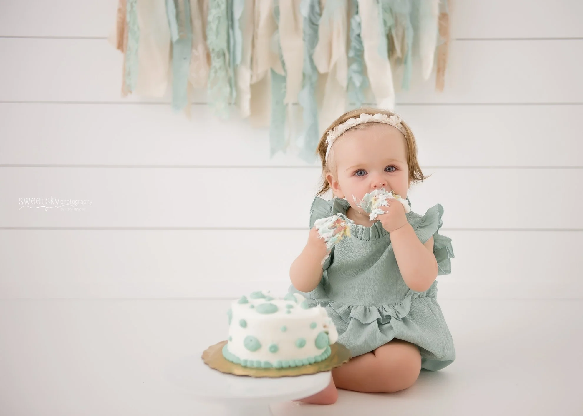 A young girl with a cream headband and a ruffled dress, sitting on the floor, eating cake with frosting on her face, with a birthday cake on a plate in front of her.