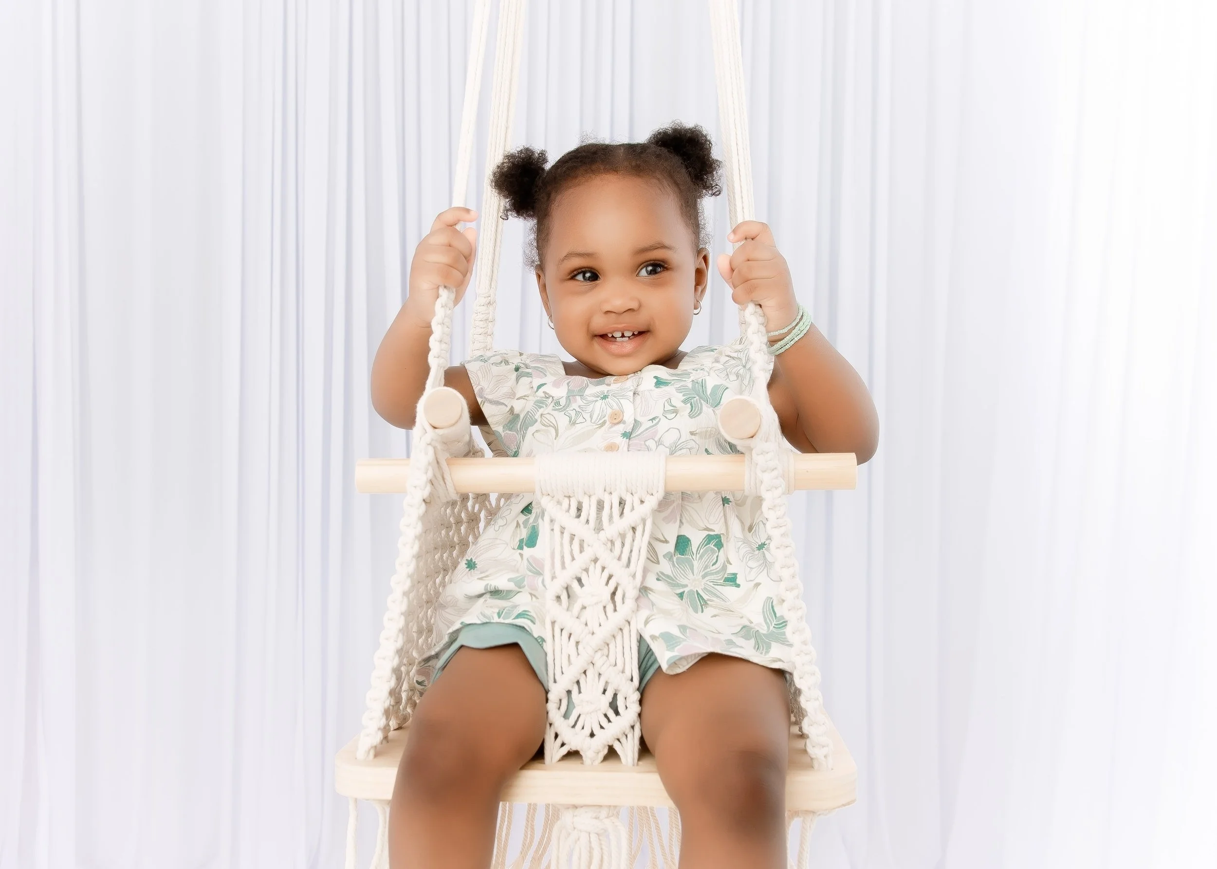 A young girl with puffy hair in pigtails sitting on a wooden swing with a woven seat and white rope, wearing a floral dress and smiling.
