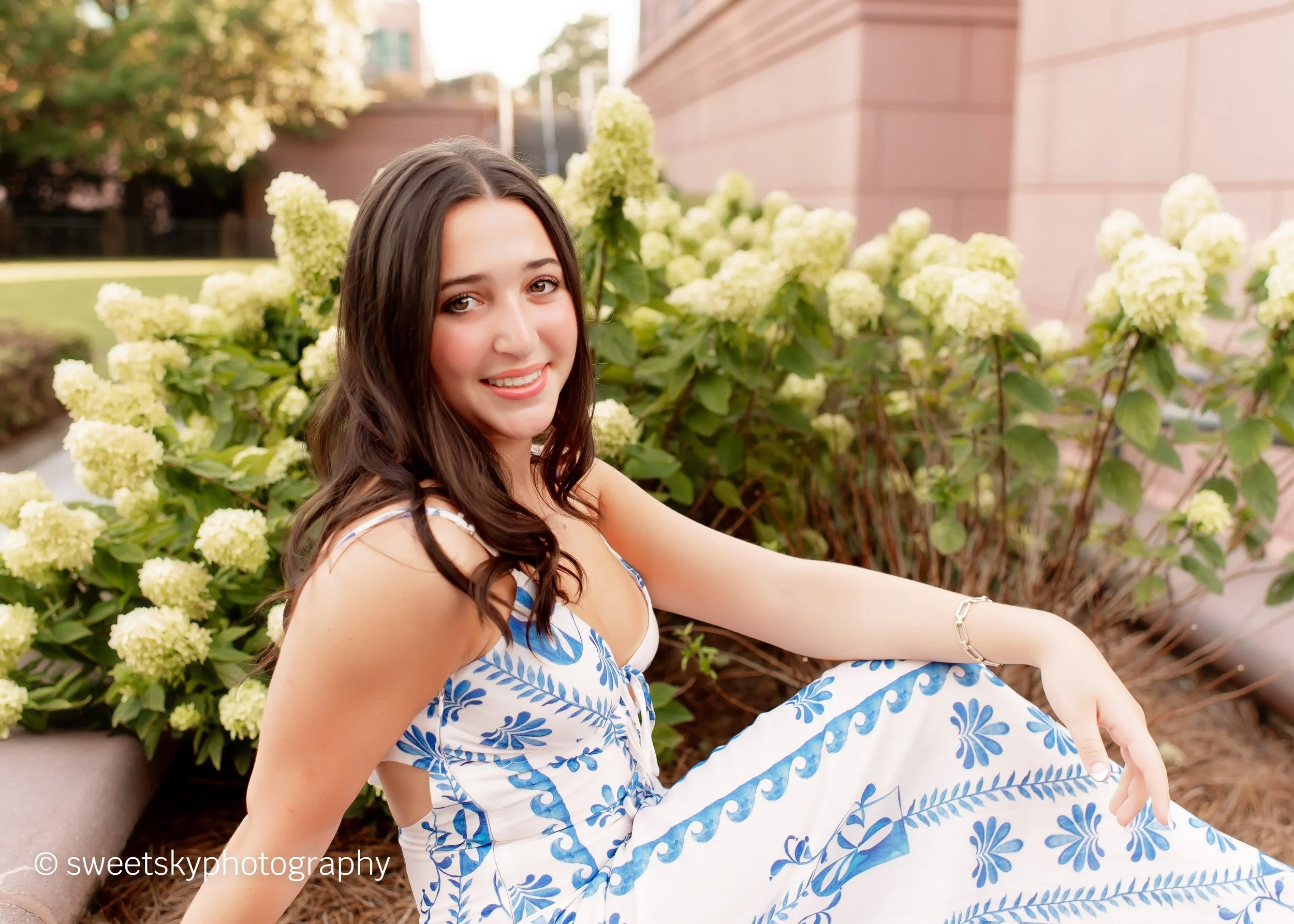 A young woman with long brown hair sitting outdoors next to a bush with white hydrangea flowers, smiling and looking at the camera, wearing a white and blue patterned dress.