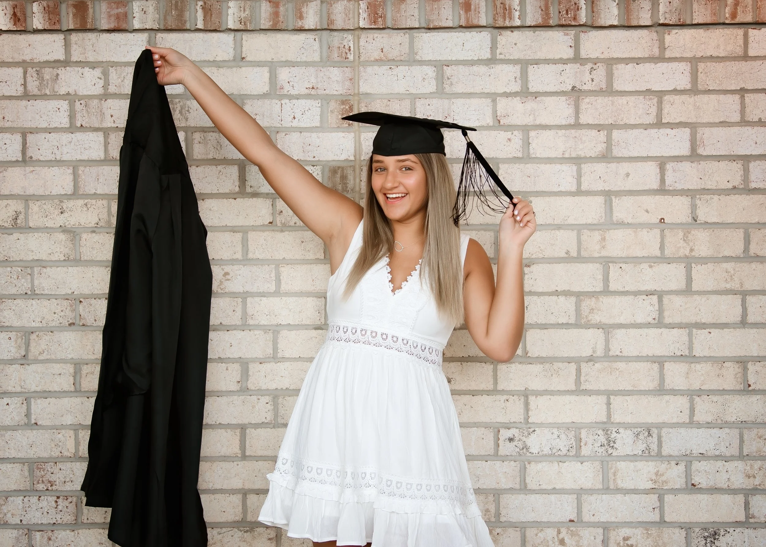 A young woman wearing a white dress and a graduation cap, smiling at the camera while holding a black gown in her left hand and adjusting her cap with her right hand, standing in front of a brick wall.