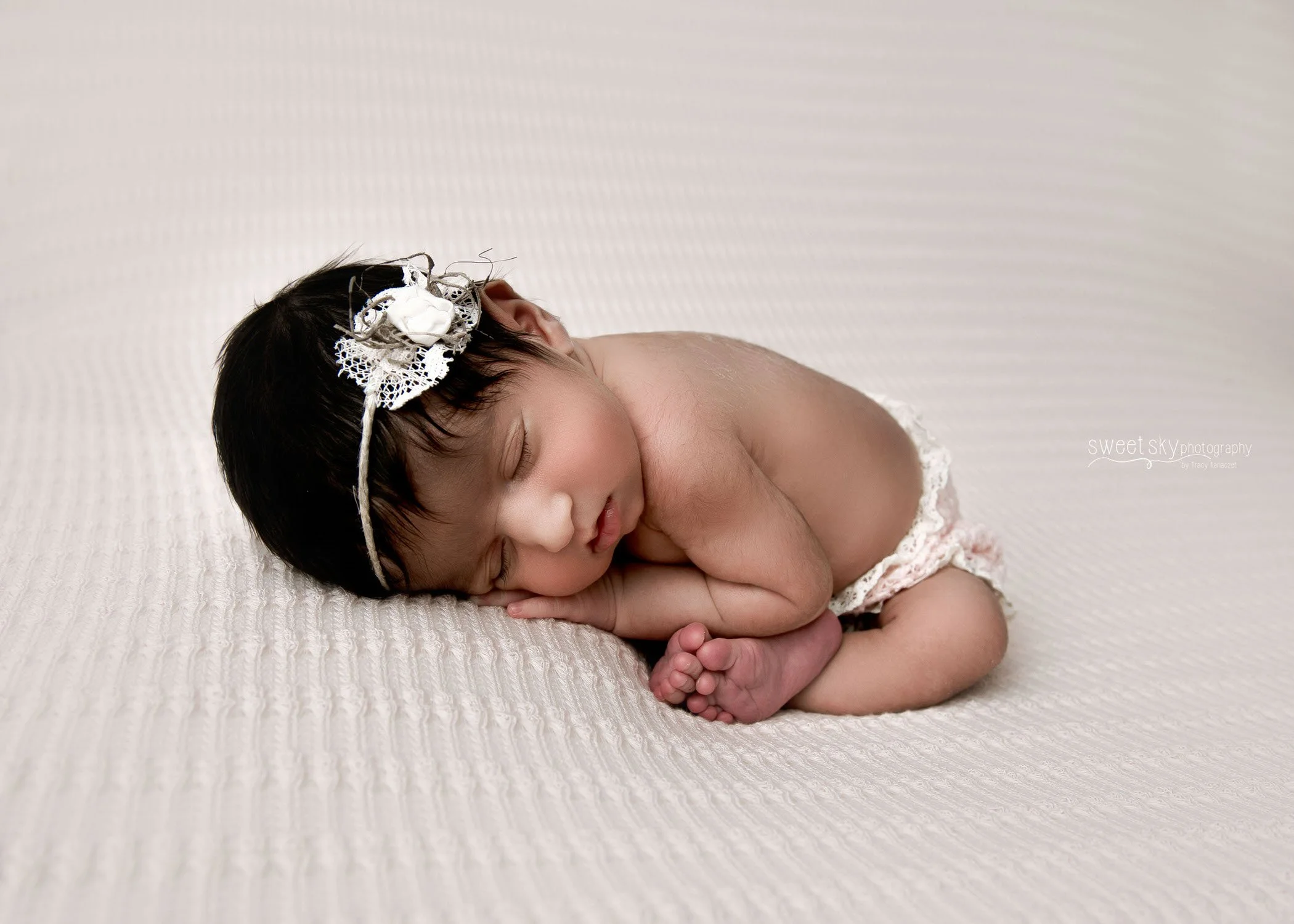 A sleeping newborn baby girl with dark hair lying on a soft, cream-colored textured blanket, wearing a white lace diaper and a decorative headband with white and gray flowers.