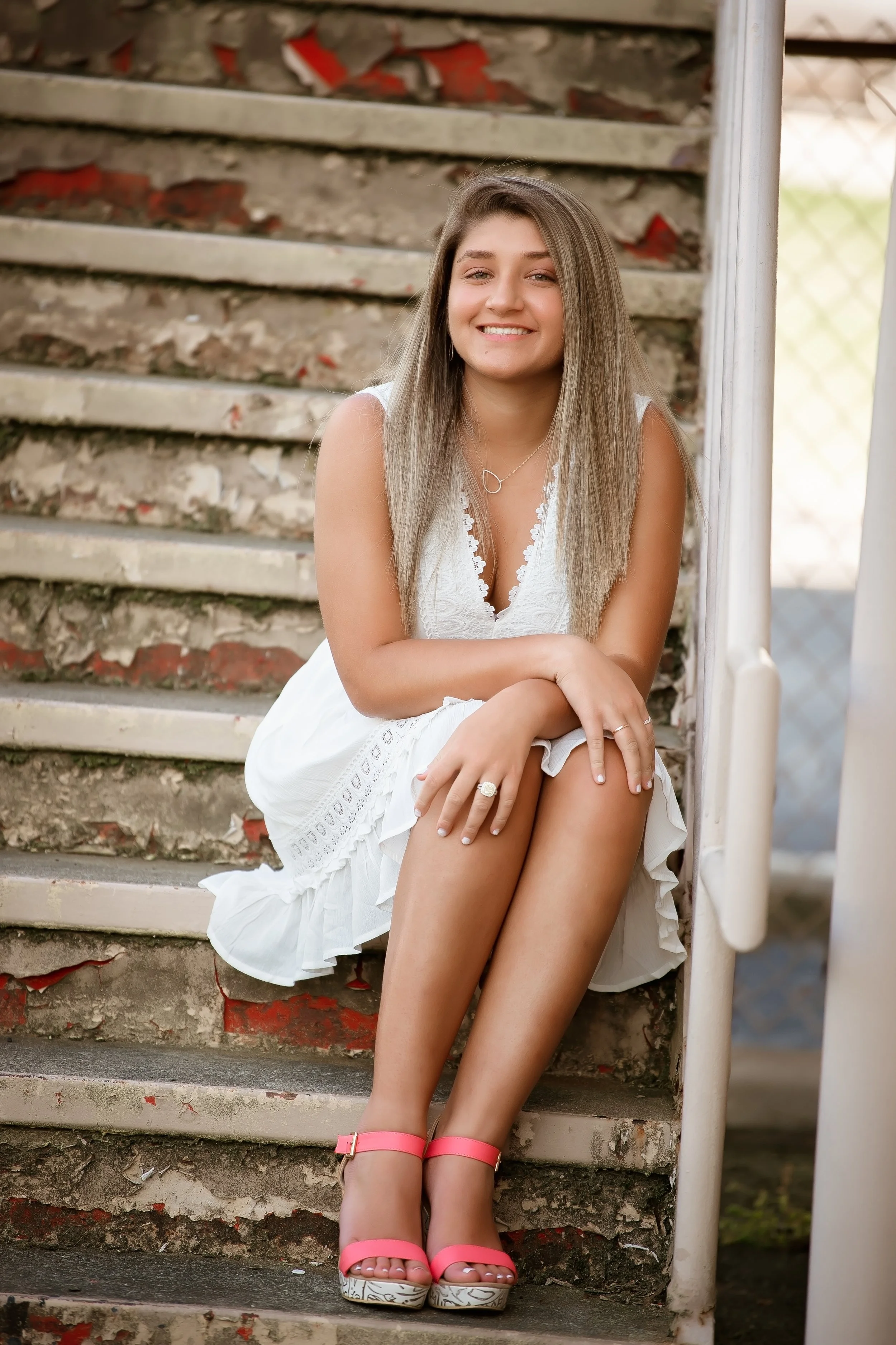 A young woman with long blonde hair sitting on concrete stairs with peeling paint, wearing a white dress and pink high-heeled sandals, smiling at the camera.